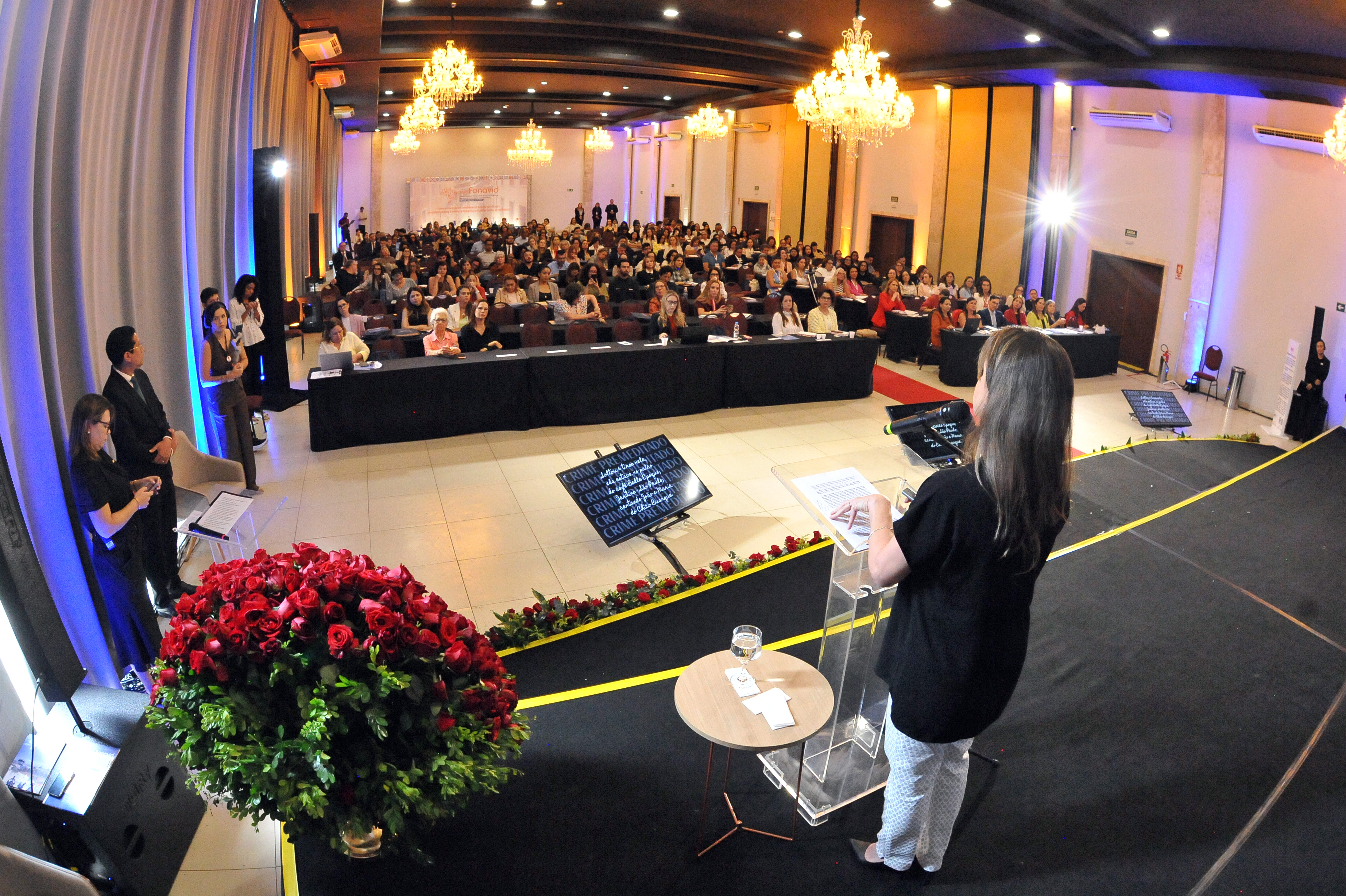 Uma foto de ângulo aberto e ligeiramente superior mostra um grande salão de eventos iluminado. No centro, uma mulher de costas para a câmera, vestindo preto e calças de padrão claro, está sobre um palco, atrás de um púlpito de acrílico. O palco é preto e decorado com um grande arranjo esférico de rosas vermelhas à esquerda. Centenas de pessoas estão sentadas no auditório, à frente do palco, acompanhando o evento.