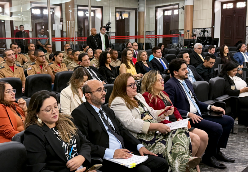 Foto horizontal da eleição de lista tríplice de advogados para o TRE. A foto exibe uma plateia em uma sessão do TJMA. Vários indivíduos estão sentados e atentos, voltados para a frente (direção onde a foto está sendo tirada). Muitos estão com trajes sociais (ternos, blazers), e alguns seguram documentos ou olham para seus celulares. Há um grupo significativo de pessoas vestindo uniformes militares de cor cáqui. O salão tem piso claro e paredes com portas ou janelas altas em madeira escura. Grandes pilares de sustentação são visíveis.