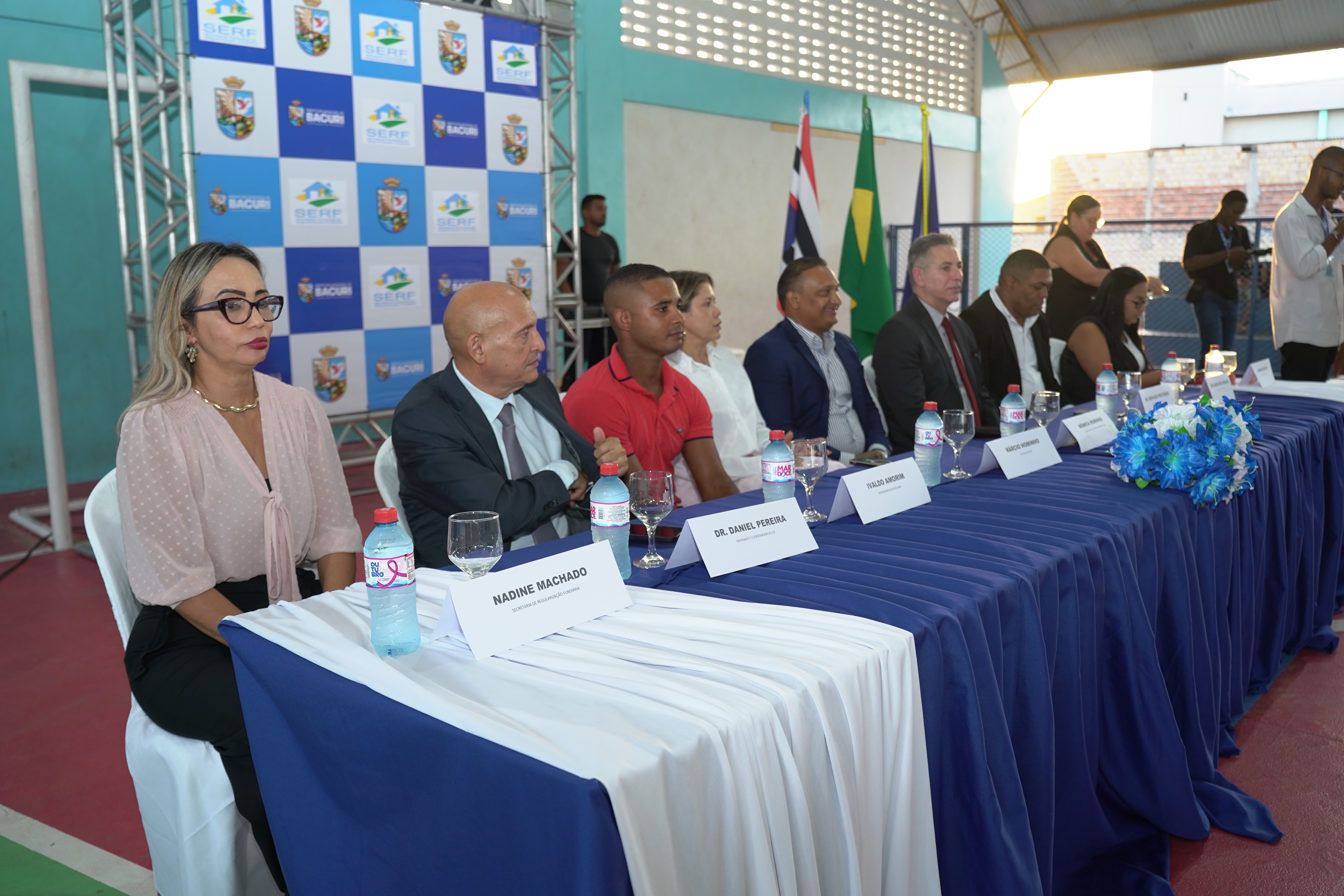 A imagem mostra a mesa de honra de um evento realizado em um local que parece ser uma quadra esportiva coberta, com estrutura metálica e paredes de tijolos vazados pintados de azul claro no fundo. A luz natural entra pela lateral direita. Primeiro Plano: A mesa é longa, coberta por toalhas em azul marinho e branco. Sobre a mesa, há garrafas de água, copos e um arranjo floral pequeno com flores azuis. Várias pessoas estão sentadas lado a lado.