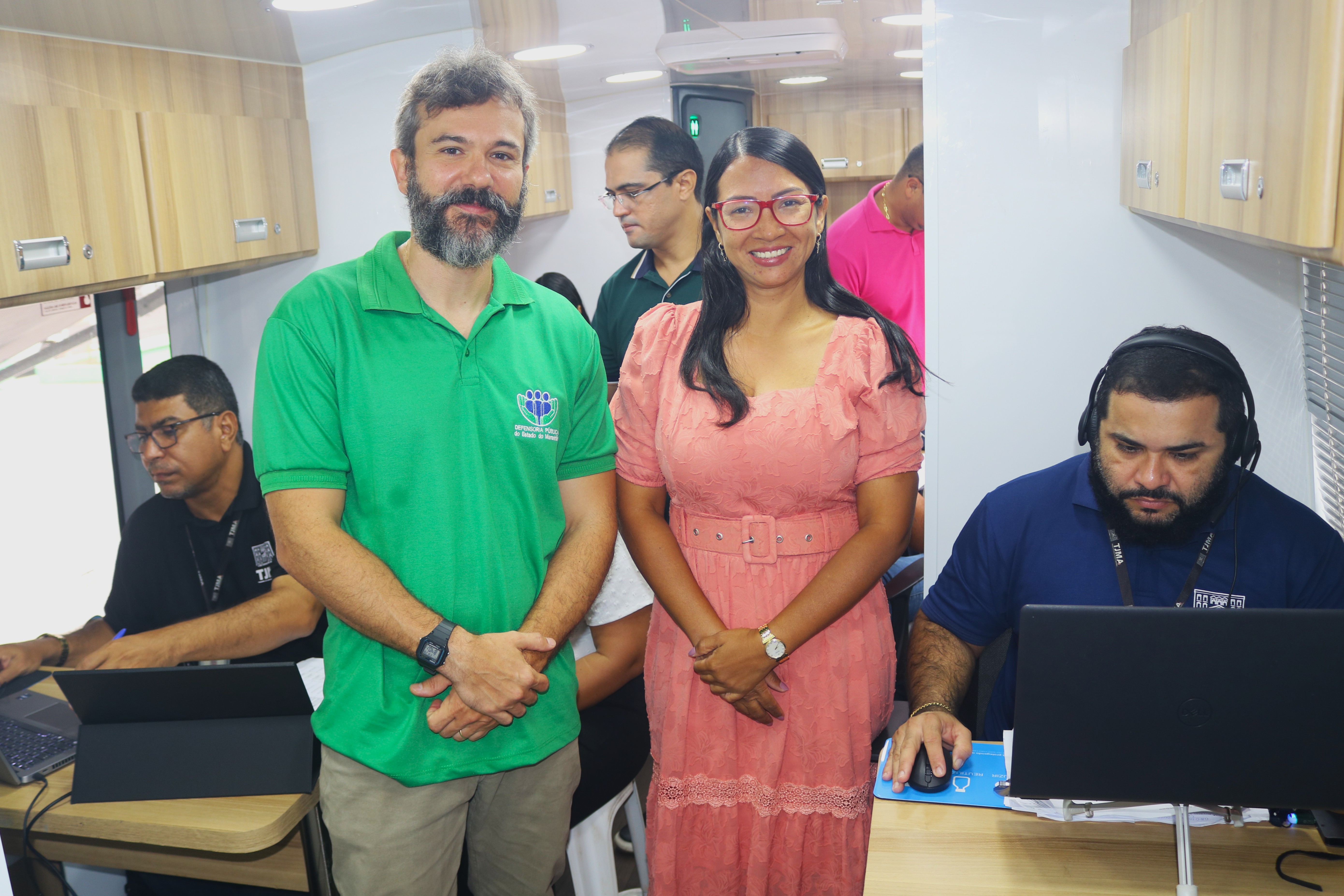 ## ¿ Descrição da Imagem  A imagem mostra o interior do veículo (o ônibus de atendimento) que está sendo utilizado para a ação de cidadania, focando em dois profissionais em destaque e no ambiente de trabalho organizado.  **Pessoas em Destaque (Centro):**  1.  **Homem (Esquerda):** Veste uma camisa polo verde brilhante, com um logotipo (provavelmente relacionado ao órgão promotor) no peito. Ele tem barba e cabelo grisalhos, e usa um relógio de pulso escuro. Ele está de pé, olhando diretamente para a câmera, com as mãos cruzadas à frente. 2.  **Mulher (Direita):** Veste um vestido salmão ou rosa claro com mangas bufantes e um cinto da mesma cor. Ela usa óculos com armação vermelha chamativa e está sorrindo para a câmera.  **Ambiente e Outros Funcionários:**  * **Configuração do Ônibus:** O interior do veículo é adaptado para ser um escritório móvel. Possui armários superiores de madeira clara com portas e puxadores metálicos, dando um aspecto de consultório ou escritório. O teto tem iluminação embutida. * **Postos de Trabalho:**     * **Esquerda:** Um homem de camisa preta está sentado em uma mesa/balcão, trabalhando em um laptop e uma tela destacável (tablet).     * **Direita:** Um homem de camisa polo azul escura está sentado e concentrado, trabalhando em um laptop. Ele usa fones de ouvido (headset) grandes, sugerindo que está em comunicação ou focado em uma tarefa que exige áudio. * **Fundo:** Outros funcionários ou pessoas em atendimento são visíveis no fundo: um homem de camisa escura e óculos no centro, e outro homem de camisa rosa na extrema direita.  **Em resumo, a foto ilustra o ambiente interno, climatizado e funcional, do ônibus de cidadania, mostrando os profissionais (provavelmente servidores do TJMA ou parceiros) prontos para o atendimento, com a dupla central posando para a foto.**  ---  Gostaria de ver uma descrição de outra imagem ou de pesquisar sobre o contexto e a localização desta ação de cidadania do TJMA?