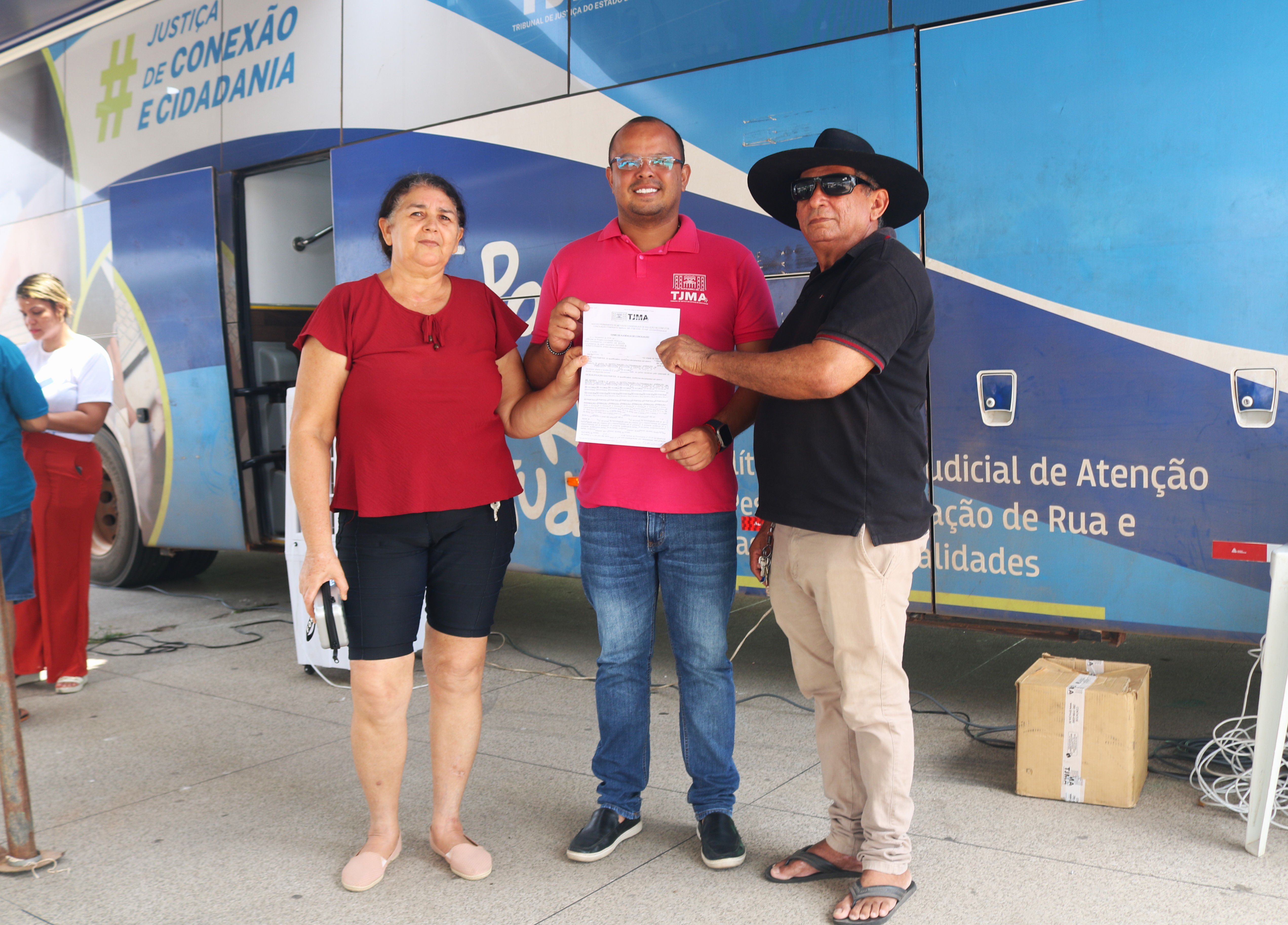 ## ¿ Descrição da Imagem  A imagem mostra três pessoas em frente a um grande veículo, provavelmente o mesmo ônibus visto na foto anterior, que foi adaptado para prestar serviços públicos. A foto tem foco no grupo central e sugere a finalização de um atendimento ou a entrega de um documento.  **Pessoas em Destaque:**  1.  **Mulher (Esquerda):** Veste uma blusa vermelha simples e shorts escuros. Ela tem cabelo escuro, está segurando algo (possivelmente um celular) na mão esquerda e olha diretamente para a câmera. 2.  **Homem (Centro):** Veste uma camisa polo rosa-choque ou vermelha brilhante, calças jeans escuras e sapatos sociais/tênis escuros. Ele parece ser um funcionário ou representante do serviço, pois está segurando e apresentando um documento que parece ser a razão da foto. A camisa tem um logotipo que parece ser do **TJMA** (Tribunal de Justiça do Maranhão), confirmando a natureza pública e legal do serviço. 3.  **Homem (Direita):** Veste uma camisa polo preta, calças cáqui e chinelos. Ele usa um chapéu preto de abas largas e óculos de sol, dando-lhe uma aparência distinta. Ele está recebendo ou manuseando o documento junto com o homem do centro.  **Plano de Fundo (O Ônibus de Serviço):**  * Atrás das pessoas, está o lado de um ônibus grande, de cor predominantemente azul e branca. * No corpo do ônibus, são visíveis várias inscrições em português, destacando a natureza do serviço:     * No canto superior esquerdo: **"JUSTIÇA DE CONEXÃO E CIDADANIA"**.     * Abaixo, na porta (ou perto dela): **"TRIBUNAL DE JUSTIÇA DO ESTADO DO..."** (o resto está cortado, mas o TJMA na camisa sugere *Maranhão*).     * No centro, um grande logotipo com a sigla **TJMA**.     * Na parte inferior, à direita: **"Judicial de Atenção ção de Rua e Cidades"** (a visibilidade está parcial, mas indica um serviço de atenção a populações específicas). * À esquerda, parcialmente visível, está outra mulher em pé, com calças vermelhas e uma camiseta branca. * Um objeto de papelão (caixa) está no chão à direita.  **Em resumo, a imagem retrata um momento de entrega de documentos em um evento de cidadania promovido pelo Tribunal de Justiça do Maranhão (TJMA), provavelmente parte de uma ação itinerante.**  ---  Posso procurar mais informações sobre a ação **"Justiça de Conexão e Cidadania"** ou o projeto móvel do **TJMA** se desejar!