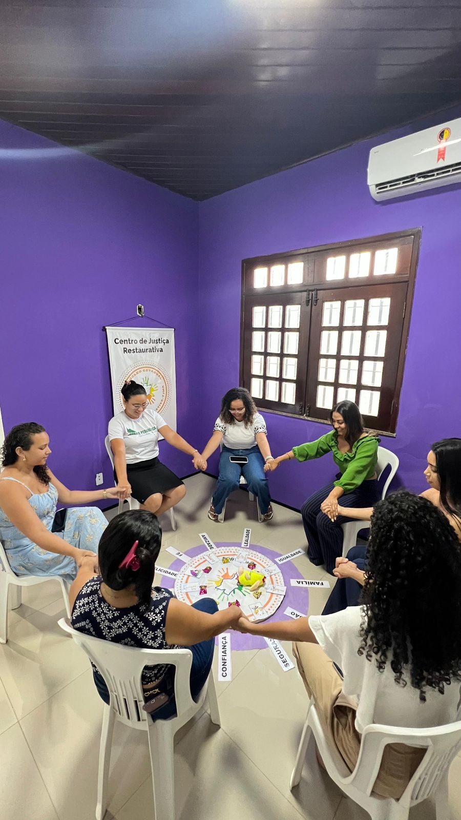 em ambiente interno, um grupo de pessoas entre homens e mulheres em pé de maos dadas em formato circular, participam de atividade com objetos dispostos no chao no centro da ação