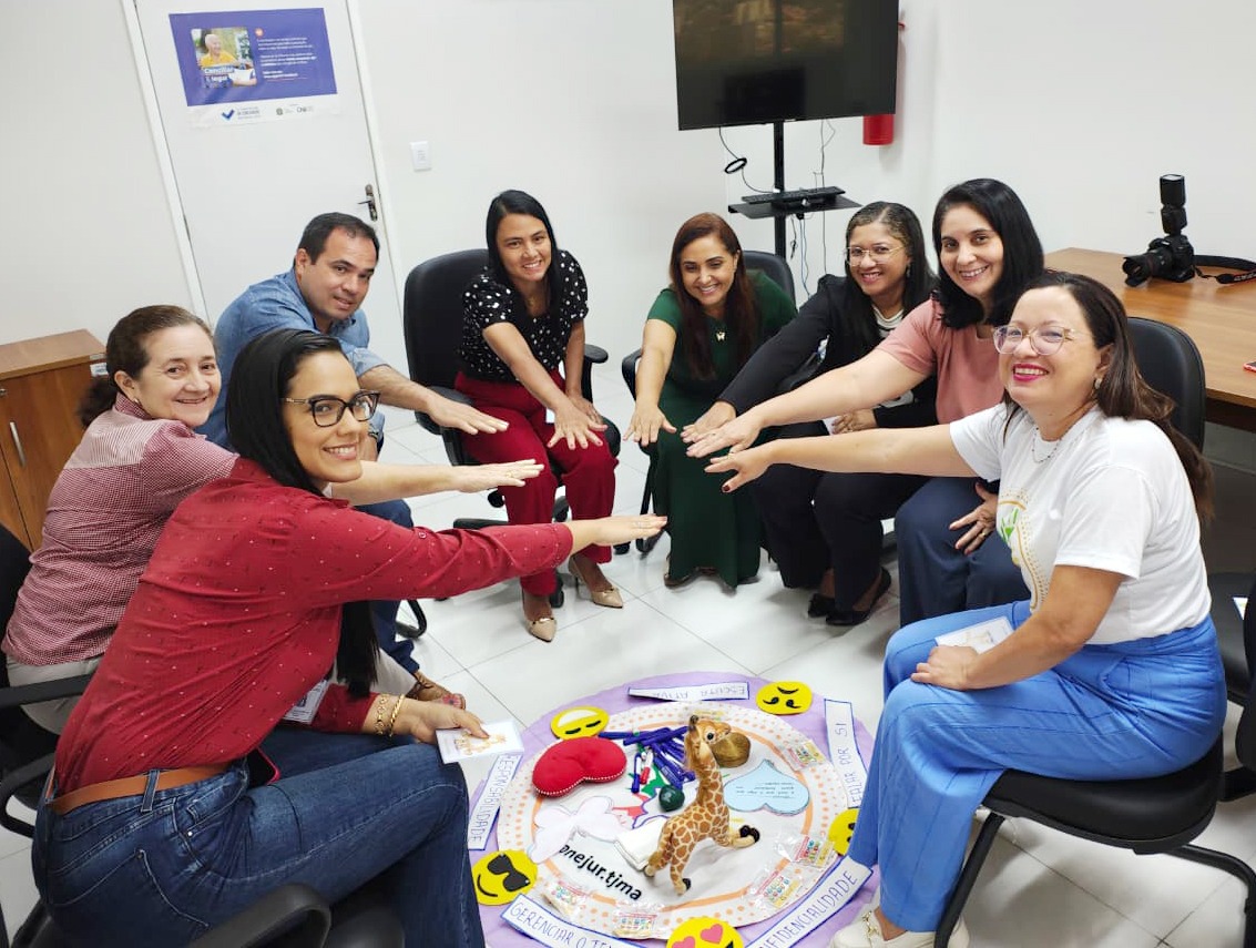 A imagem mostra um grupo de oito pessoas (sete mulheres e um homem) reunidas em uma sala de escritório, sentadas em cadeiras de rodinhas e dispostas em círculo. O ambiente parece ser de trabalho ou treinamento, com uma TV em um suporte no fundo e cartazes na parede.

No centro do círculo, há um grande painel redondo no chão, colorido e decorado com emojis, notas musicais, corações e um brinquedo de girafa. Os participantes esticam os braços para o centro, com as mãos sobrepostas sobre o painel, em um gesto que sugere união, colaboração ou um acordo/início de atividade. Todos na foto parecem estar sorrindo e engajados.