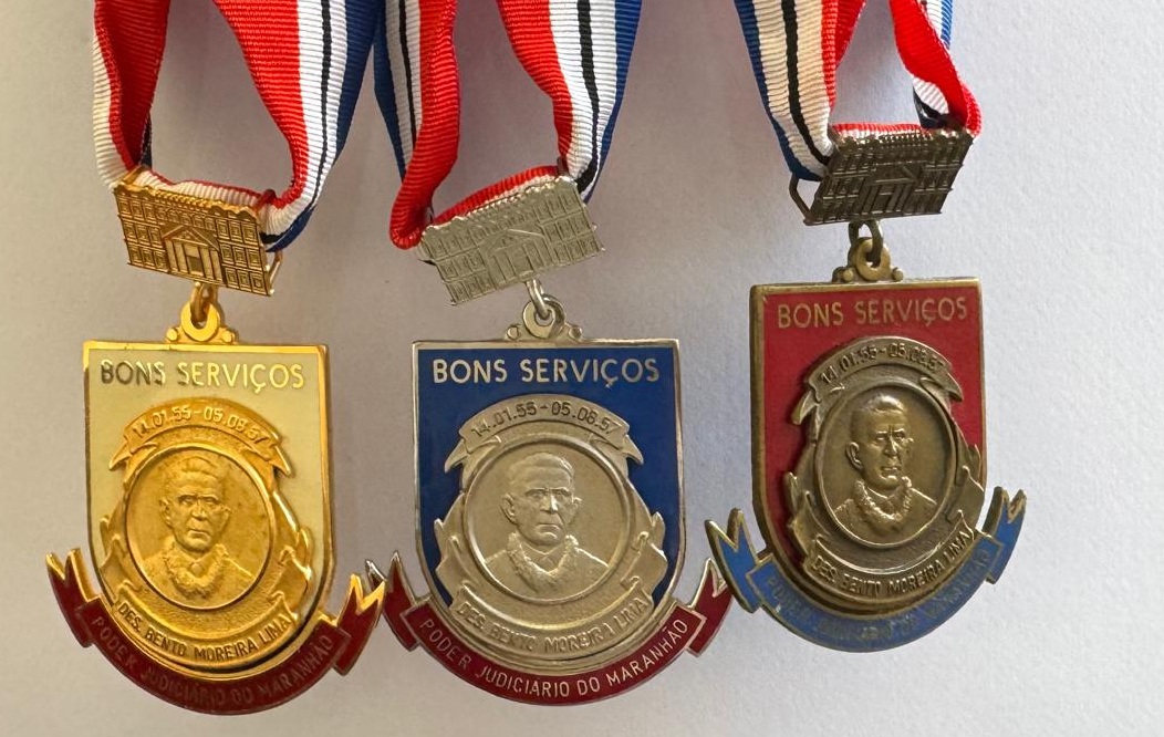 Foto horizontal de medalhas Bento Moreira Lima. A imagem mostra três medalhas de premiação ou condecoração: uma é dourada com detalhes em vermelho, uma é prateada com fundo azul escuro e detalhes em vermelho, e a terceira é em um metal de cor cobre/bronze com fundo vermelho e detalhes em azul. Todas estão suspensas por uma fita com as cores vermelho, branco e azul em listras verticais. O suporte da fita que se conecta à medalha tem a forma de um pequeno edifício. No topo, está escrito: BONS SERVIÇOS. No centro do medalhão: a efígie (rosto) do Desembargador Bento Moreira Lima. Nas faixas na parte inferior: DES. BENTO MOREIRA LIMA e PODER JUDICIÁRIO DO MARANHÃO.
