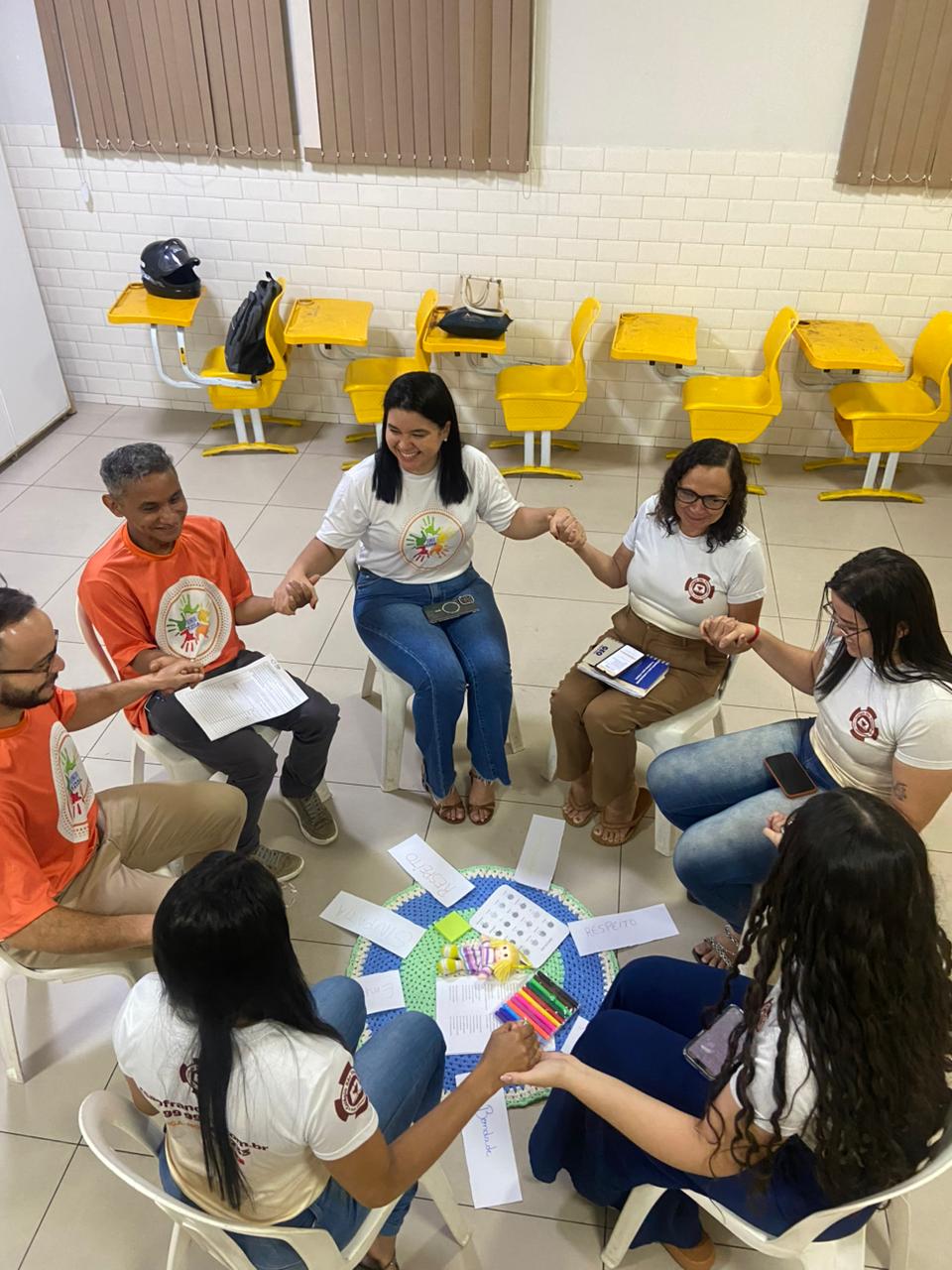 em ambiente interno, um grupo de pessoas entre homens e mulheres sentados e dispostos em formato circular, participam de atividade com objetos dispostos no chao no centro da ação