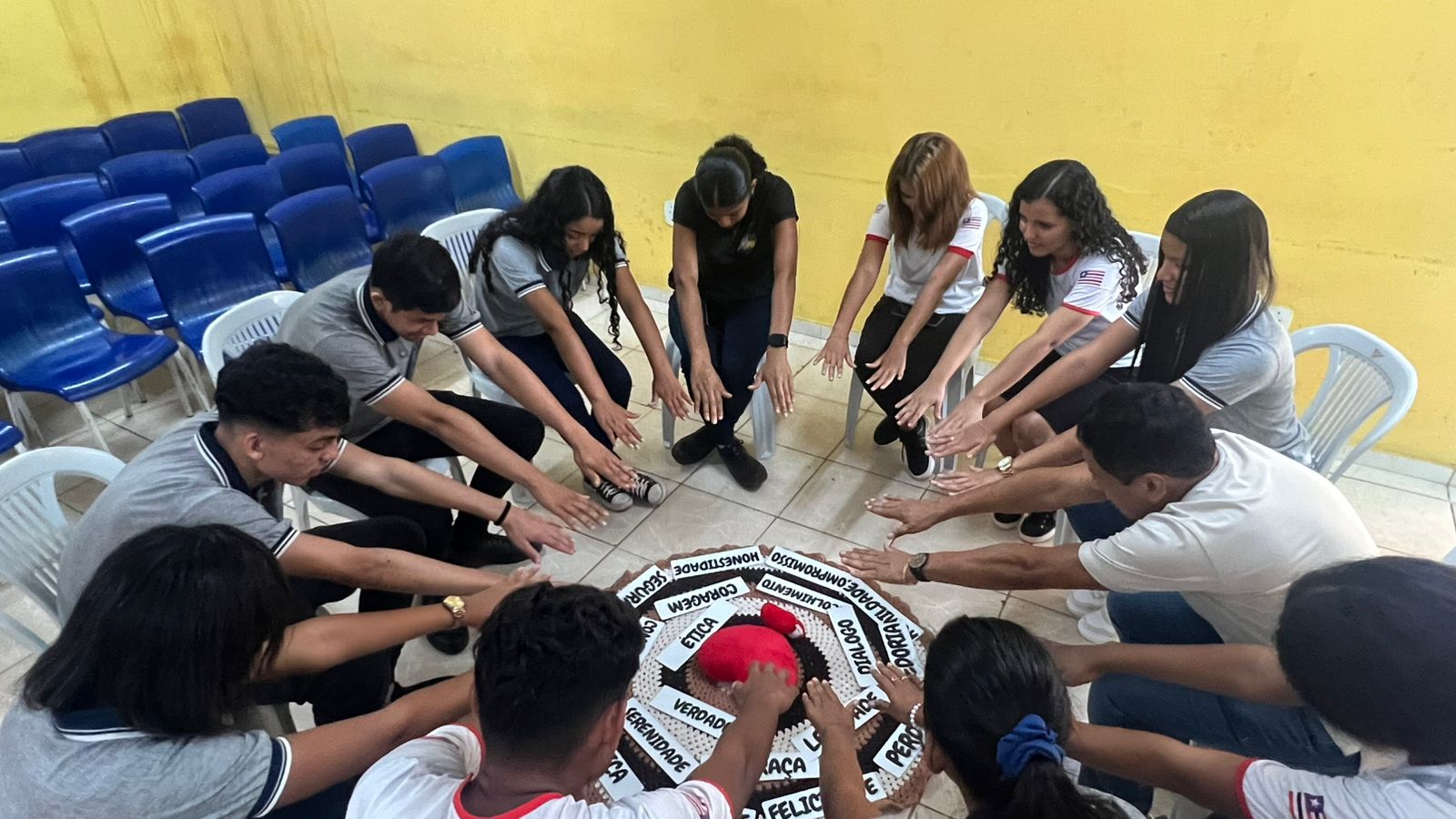 em ambiente interno, um grupo de pessoas entre homens e mulheres sentados e dispostos em formato circular, participam de atividade com objetos dispostos no chao no centro da ação