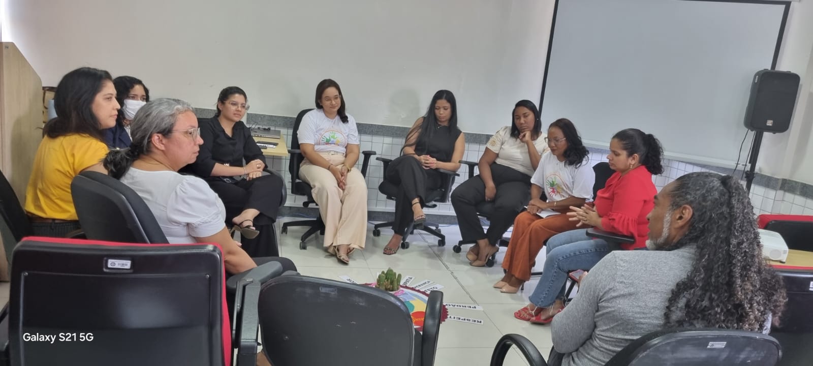 em ambiente interno, grupo de pessoas sentadas em formato cirular participam de atividade