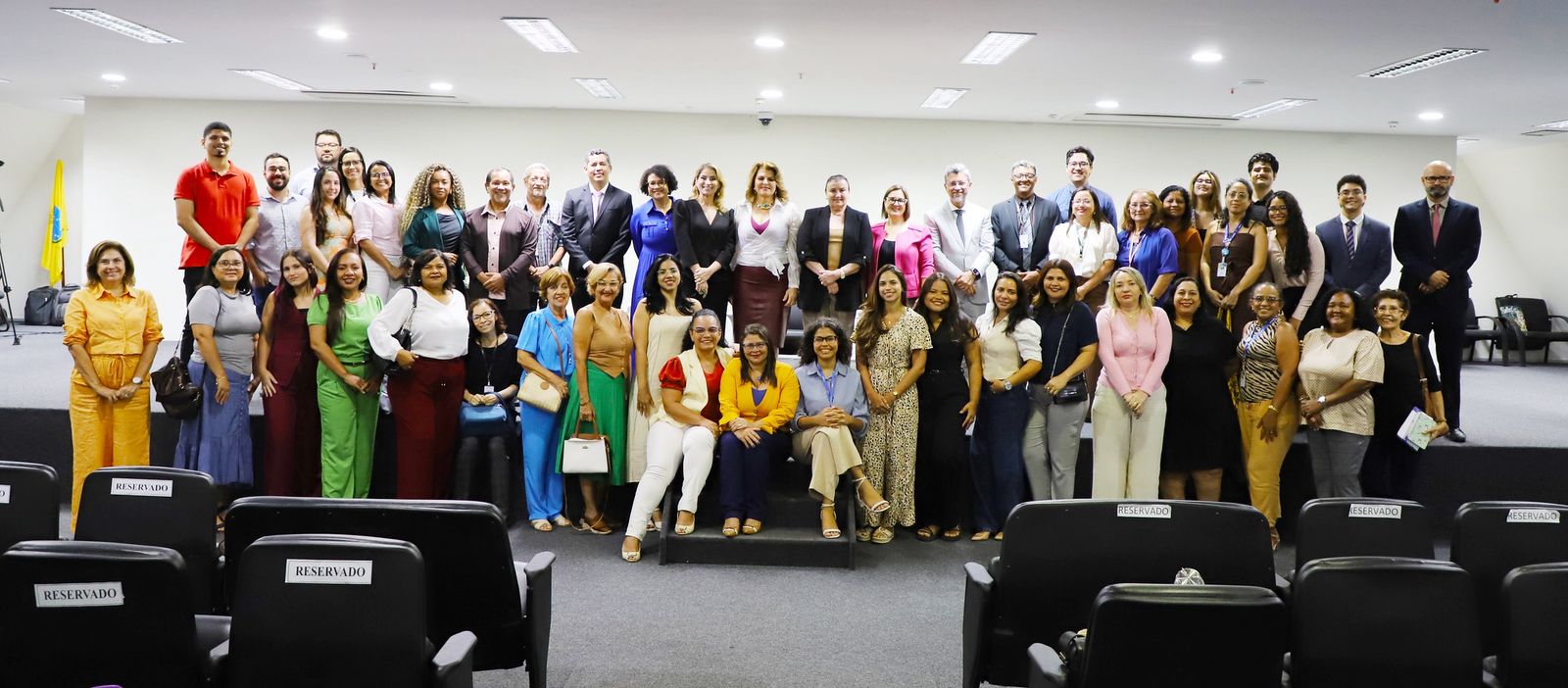 A imagem mostra um grande grupo de pessoas, aproximadamente quarenta e cinco participantes, reunidas em frente ao palco de um auditório do TJMA. Estão em duas fileiras: a primeira sentada nos degraus, a segunda em pé. Há diversidade de idade, gênero e vestimentas. Ao fundo, é possível ver a parede clara do auditório, cadeiras e, à esquerda, uma bandeira institucional. A câmera está posicionada a partir das cadeiras reservadas da plateia.