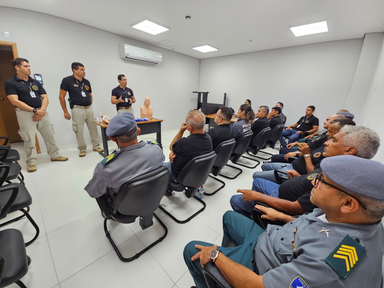 Imagem registra uma sala de treinamento, com agentes de segurança sentados em cadeiras pretas, voltados para três instrutores posicionados à frente da sala. Sobre a mesa central há materiais de instrução, incluindo um boneco utilizado para demonstração de primeiros socorros. Os participantes, entre policiais militares e agentes do TJMA, acompanham a explicação do instrutor, em ambiente iluminado e organizado.
