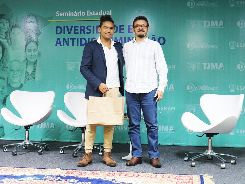 Foto colorida mostra o facilitador gráfico Sidan ORafa em pé ao lado do designer da Ascom do TJMA, Carlos Eduardo Sales, segurando uma sacola, durante palestra, no Seminário de Diversidade e Antidiscriminação, no Fórum de São Luís.