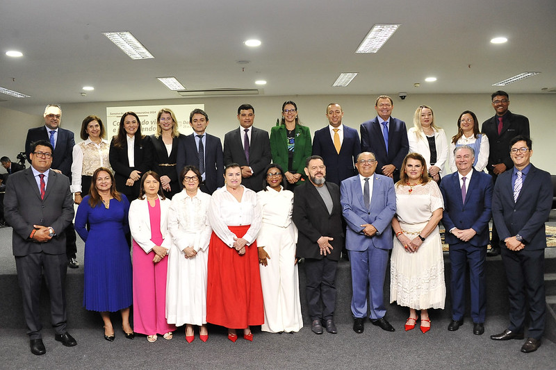 Foto horizontal de integrantes da abertura da Semana da Justiça Restaurativa. A imagem mostra um grupo de pessoas posando para uma foto em um ambiente interna. Há três fileiras de pessoas: a fileira da frente tem nove pessoas, a fileira do meio tem seis pessoas e a fileira de trás tem seis pessoas. As pessoas estão vestidas formalmente, com ternos, vestidos e roupas sociais. Ao fundo, há uma tela com um letreiro, mas não está totalmente legível na imagem. O ambiente é bem iluminado com luzes de teto.