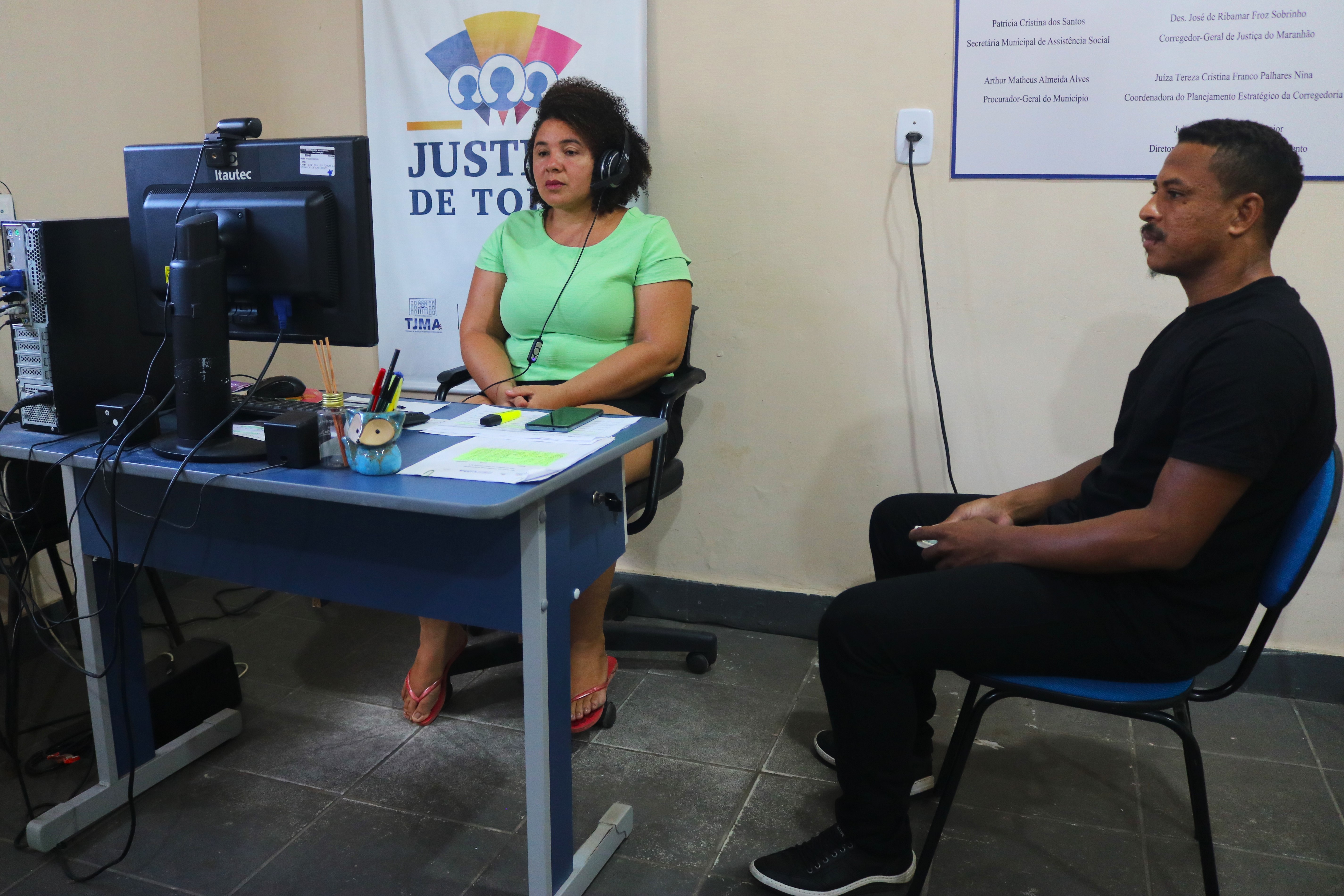 A imagem mostra uma pessoa sentada em uma cadeira, usando um headset, participando de uma videoconferência em um computador. A tela do computador exibe duas pessoas em uma chamada de vídeo. A pessoa está em um ambiente de escritório, com uma mesa que contém um teclado, um mouse, um monitor, alguns papéis, um marcador de texto amarelo e outros itens de escritório. Há também um roteador e outros dispositivos eletrônicos na parede ao fundo.

Parece ser um ambiente de trabalho moderno, onde a tecnologia facilita a comunicação e a colaboração à distância.