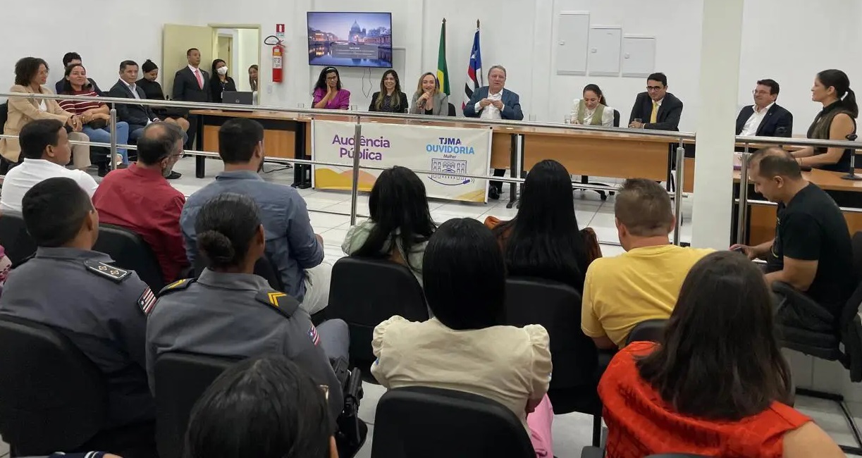 Foto horizontal de audiência pública da Ouvidoria da Mulher em Pinheiro. A imagem mostra várias pessoas sentadas em cadeiras, assistindo a uma mesa composta por várias autoridades. Na mesa, há um banner com os dizeres "Audiência Pública" e "TJMA OUVIDORIA". Atrás da mesa, há uma bandeira do Brasil e uma do Maranhão. Na parede, há uma televisão exibindo uma imagem de uma cidade. Algumas pessoas estão em pé ao fundo, próximas a uma porta e a um extintor de incêndio. A audiência ocorre em um auditório.