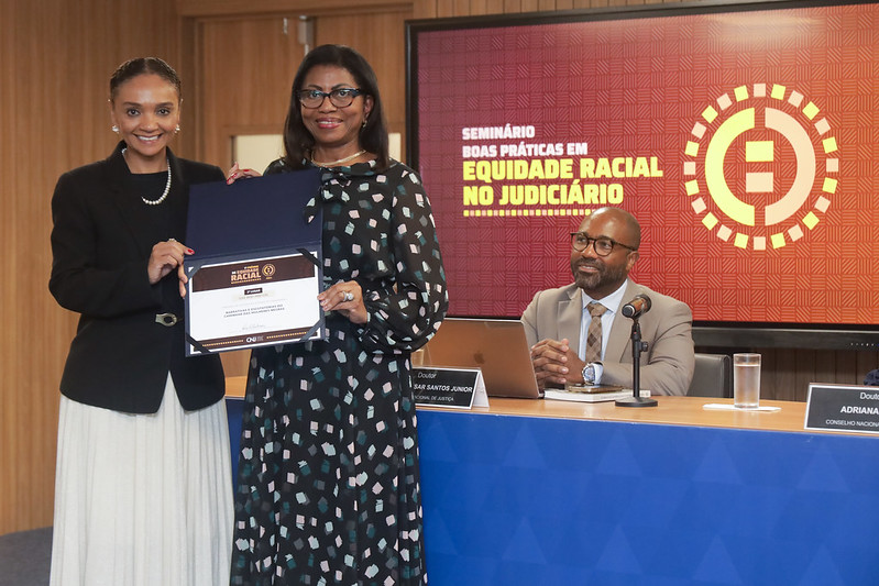 Foto horizontal do Prêmio de equidade racial - Desembargadora Angela Salazar recebe o prêmio. A imagem mostra duas pessoas em pé, uma delas, a desembargadora Angela Salazar, segurando um certificado do prêmio de terceiro lugar. Uma terceira pessoa está sentada à mesa ao fundo. A mesa possui um microfone e um laptop. No fundo, há um painel com o texto "SEMINÁRIO BOAS PRÁTICAS EM EQUIDADE RACIAL NO JUDICIÁRIO". O certificado que está sendo segurado tem o texto "EQUIDADE RACIAL NO JUDICIÁRIO" e "CERTIFICADO".