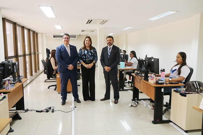 A imagem mostra um ambiente de escritório com várias pessoas trabalhando em suas mesas (é uma sala onde acontecem audiências on-line). O escritório tem várias estações de trabalho com computadores, e outros funcionários estão sentados em suas mesas trabalhando.