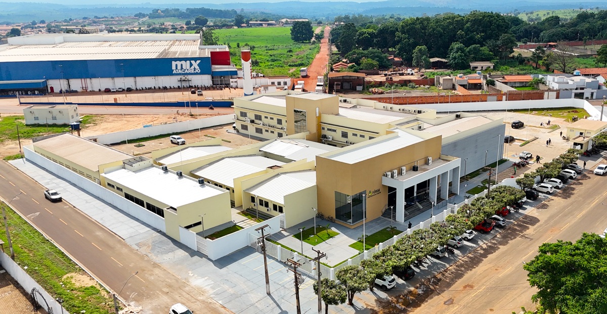 A imagem, capturada de um ponto de vista elevado (aéreo), mostra um edifício institucional ou complexo de saúde moderno, de cor amarela clara e branca, com um design de múltiplos blocos e telhados planos.

Em frente ao edifício, há um estacionamento organizado com árvores alinhadas e uma área de paisagismo gramada. A estrutura é cercada por asfalto e calçadas.

No plano de fundo e à esquerda, é visível um grande galpão industrial ou centro de distribuição azul com a inscrição "MIX". À direita e no topo da imagem, há uma área residencial e verde, com uma estrada de terra vermelha que se estende ao horizonte, indicando que o complexo está em uma área que mistura desenvolvimento urbano e paisagem natural/rural.
