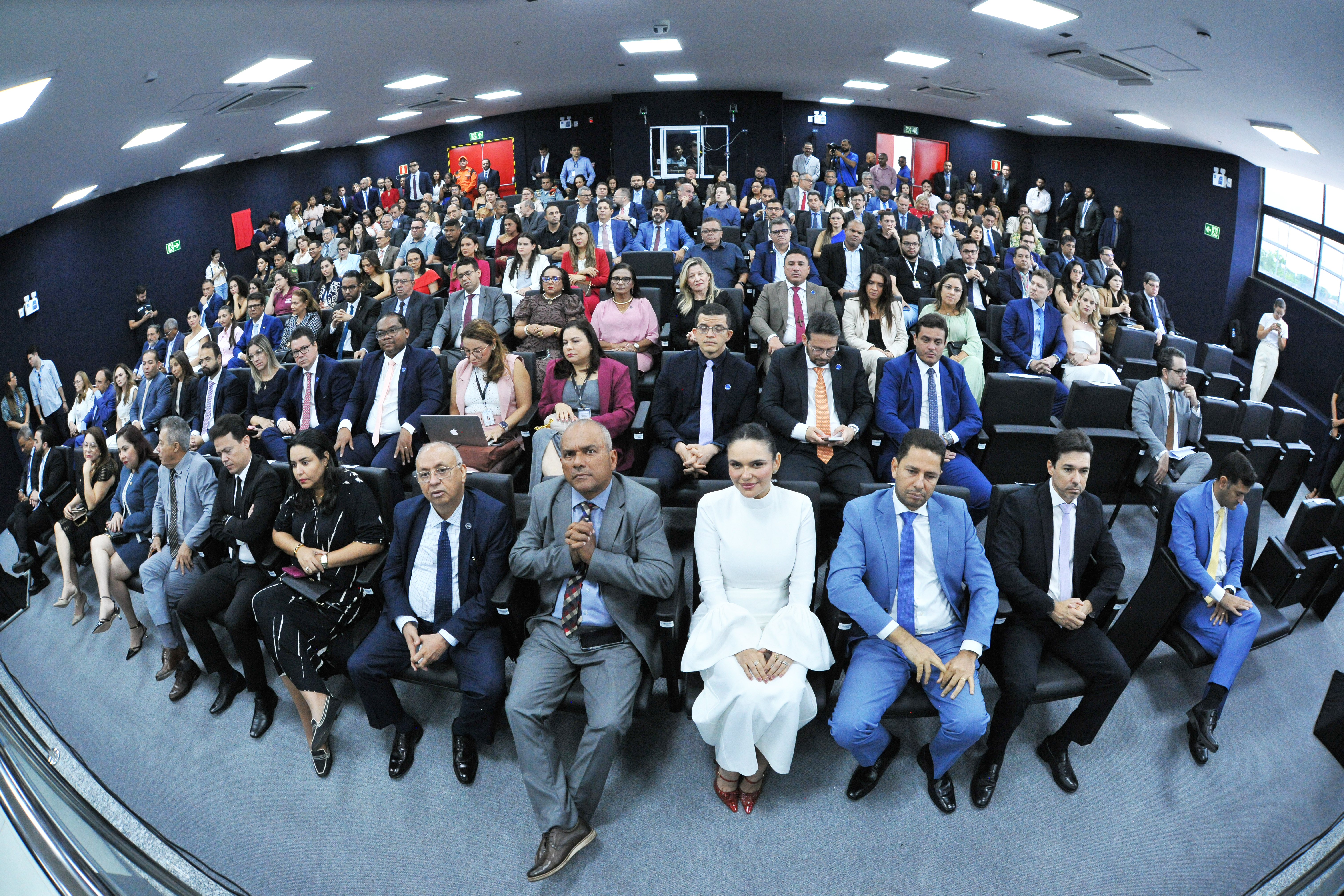 A imagem mostra cinco pessoas sentadas lado a lado em uma longa mesa, todas vestidas em trajes formais. Pela vestimenta e identificações visíveis, esta é a mesa diretora (ou mesa de honra) de um evento formal ou sessão de julgamento.