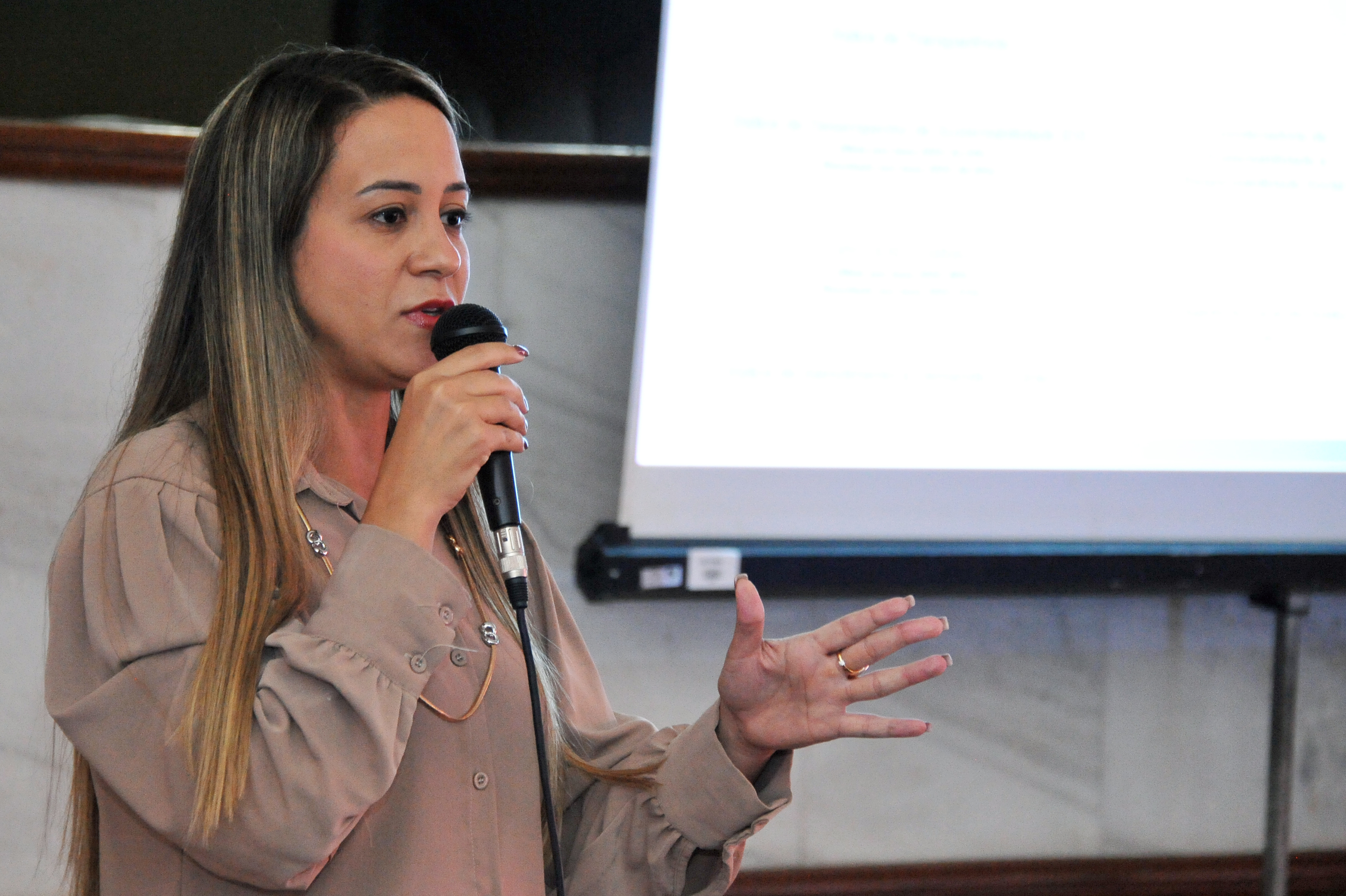 Fotografia em plano médio fechado (busto), focando na palestrante Bianca Giordano durante sua explicação.  A Palestrante: Mulher de pele clara com longos cabelos castanhos claros. Ela veste uma camisa bege.  Ação: Segura o microfone próximo à boca com a mão direita, enquanto a mão esquerda está aberta em um gesto explicativo.  Segundo Plano: Ao fundo, de forma desfocada, aparece a tela de uma projeção branca com textos e tabelas técnicas.