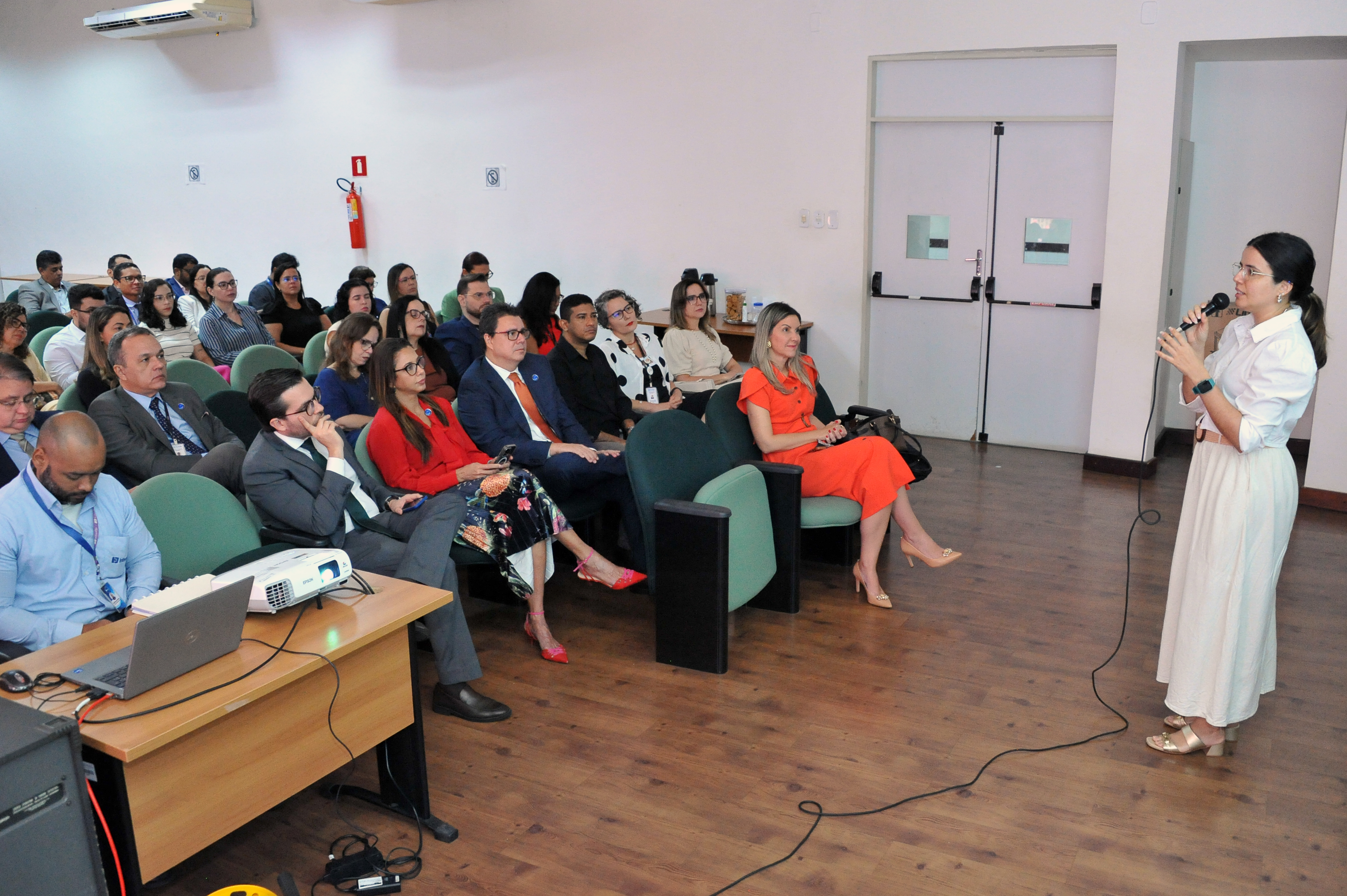 Fotografia em plano aberto mostrando a lateral do auditório durante a fala de Mariana Carneiro.  A Palestrante: À direita, uma mulher de óculos e cabelos presos em um rabo de cavalo baixo veste camisa branca social e saia longa bege. Ela segura o microfone com a mão direita.  A Plateia: A imagem captura o perfil dos participantes sentados. Na primeira fila, os gestores acompanham a fala. O foco está na profundidade do auditório, com as portas brancas ao fundo.