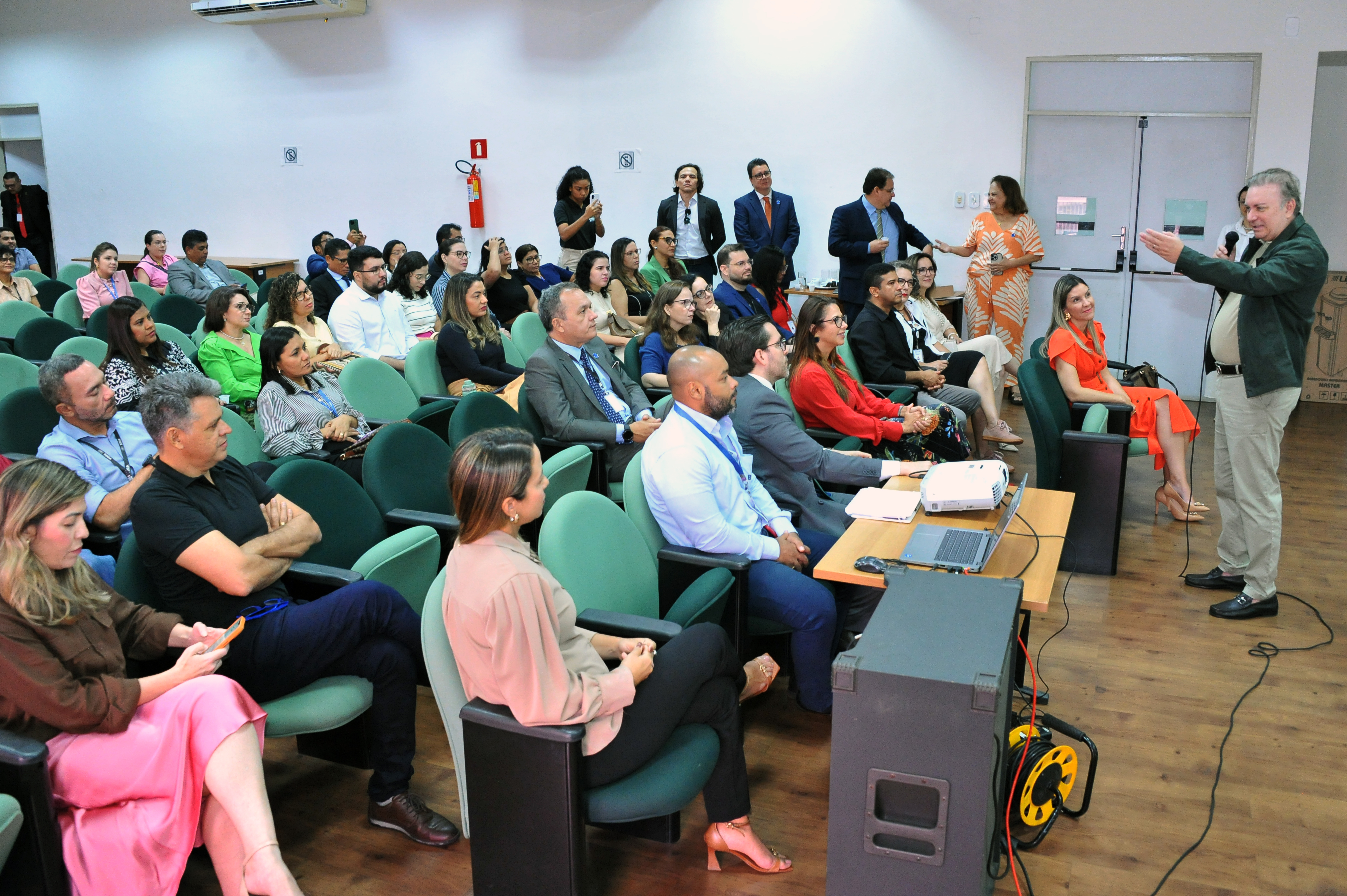 Fotografia colorida, em plano médio e ângulo levemente elevado, mostrando o interior de um auditório durante uma palestra.

O Palestrante: À direita, um homem de pele clara e cabelos grisalhos (Desembargador Froz Sobrinho) está em pé, de perfil para a esquerda. Ele veste uma jaqueta verde escura sobre uma camisa clara e calças beges. Ele segura um microfone na mão esquerda e gesticula com a direita enquanto fala para a plateia.

A Plateia: Cerca de 40 pessoas estão sentadas em poltronas estofadas de cor verde-água, dispostas em fileiras diagonais. O público é diverso, composto por homens e mulheres com roupas formais e casuais. Alguns olham atentamente para o palestrante, enquanto outros consultam celulares ou anotam informações.

O Ambiente: O auditório possui piso de madeira clara e paredes brancas. À frente da primeira fila, há uma pequena mesa de madeira com um projetor branco, um notebook aberto e alguns papéis. No canto inferior direito, vê-se parte de uma caixa de som preta e um carretel de cabos amarelos. Ao fundo, algumas pessoas permanecem em pé próximas à porta de entrada.