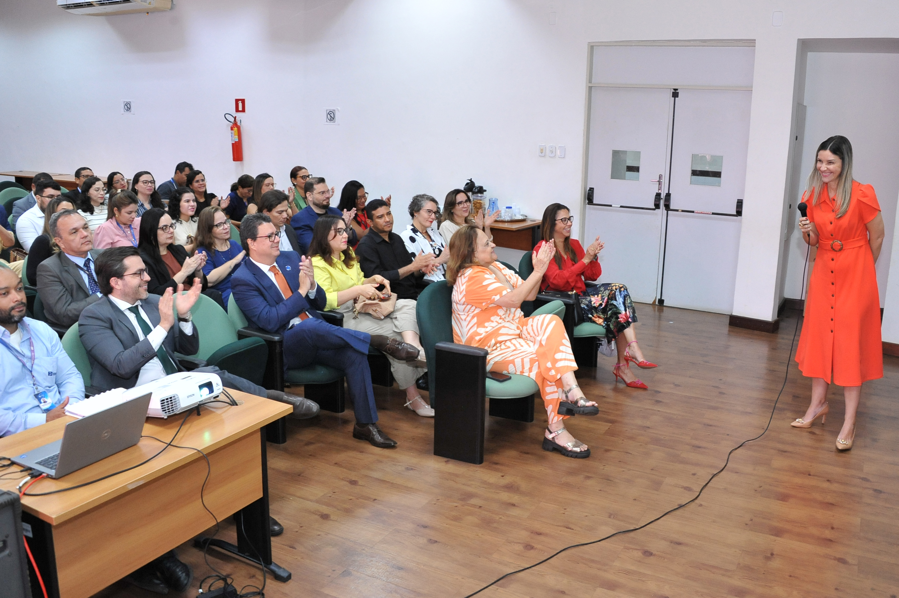 Fotografia colorida em plano aberto mostrando a plateia e a juíza Tereza Nina.  A Ação: A plateia está sentada e aplaudindo. A juíza Tereza Nina, à direita, está em pé vestindo um vestido laranja longo com botões frontais e cinto. Ela sorri para o público enquanto segura o microfone.  Ambiente: O enquadramento é similar à Imagem 1, mas o foco emocional é de celebração e encerramento.