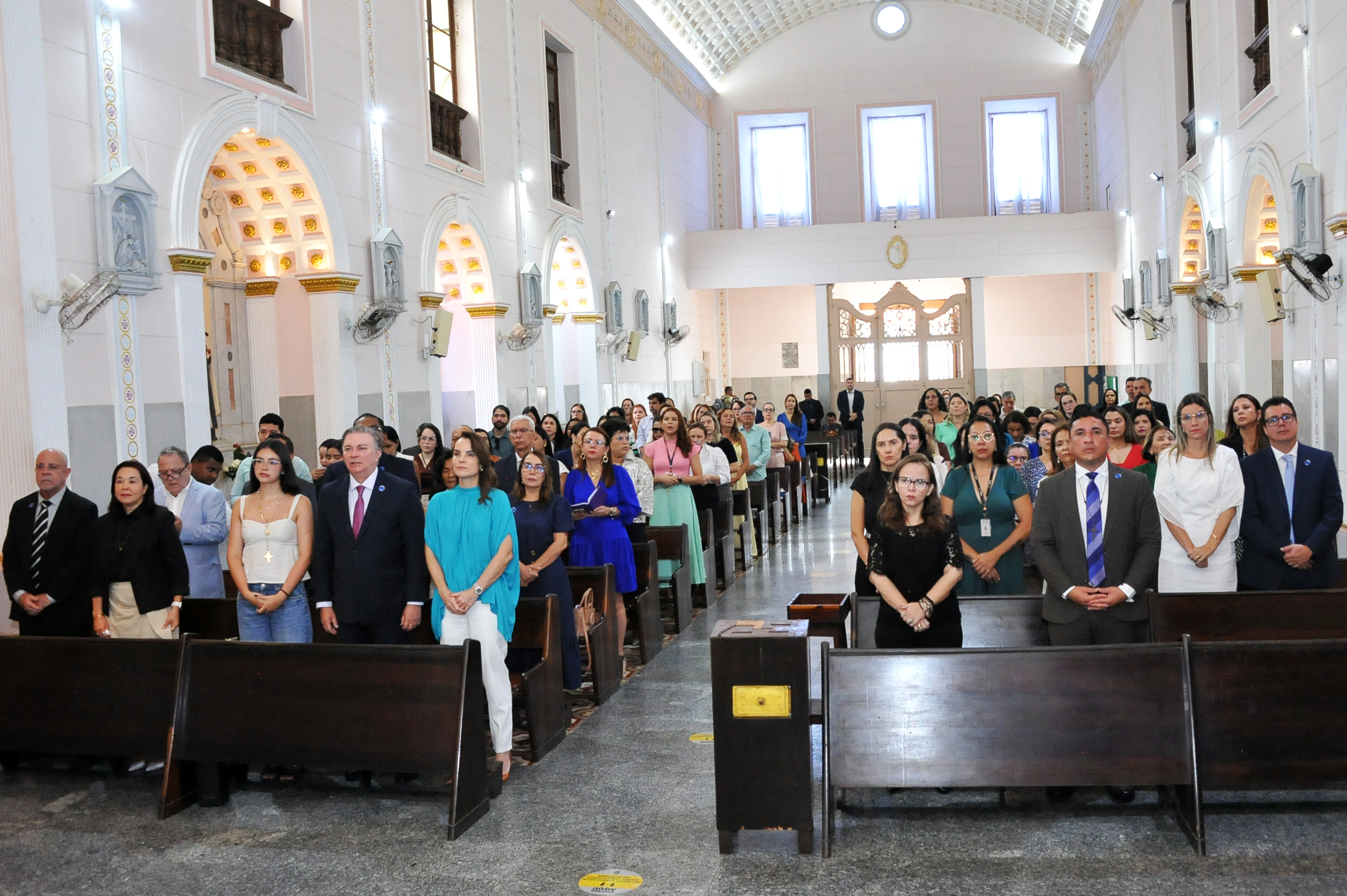 Celebração religiosa realizada no interior da Igreja de Nossa Senhora do Carmo. O espaço apresenta arquitetura clássica, com altar ornamentado, imagens sacras e iluminação clara. Um arcebispo, trajando vestes litúrgicas na cor roxa, conduz a celebração junto ao altar. Ao fundo, outros membros do clero acompanham o momento. Em outra cena, autoridades, magistrados, servidores e servidoras do Tribunal de Justiça do Maranhão estão em pé, distribuídos pelos bancos da igreja, participando da missa. Em destaque, há também um momento de fala ao púlpito, diante do altar, com o público atento, em ambiente solene e de recolhimento.