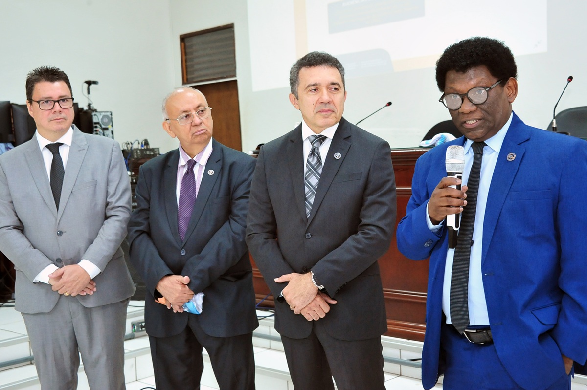 Um grupo de homens, de terno e gravata, em pé lado a lado em ambiente interno. Um dos homens aparece falando ao microfone.