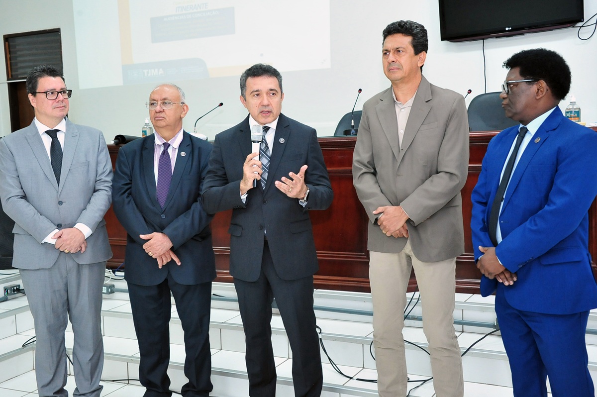 Um grupo de homens, de terno e gravata, em pé lado a lado em ambiente interno. Um dos homens aparece falando ao microfone.