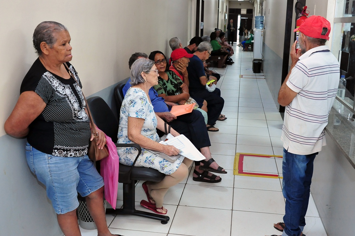 em corredor interno de corredor, pessoas aguardam atendimento sentadas e outras em pé, na maioria idosos e idosas