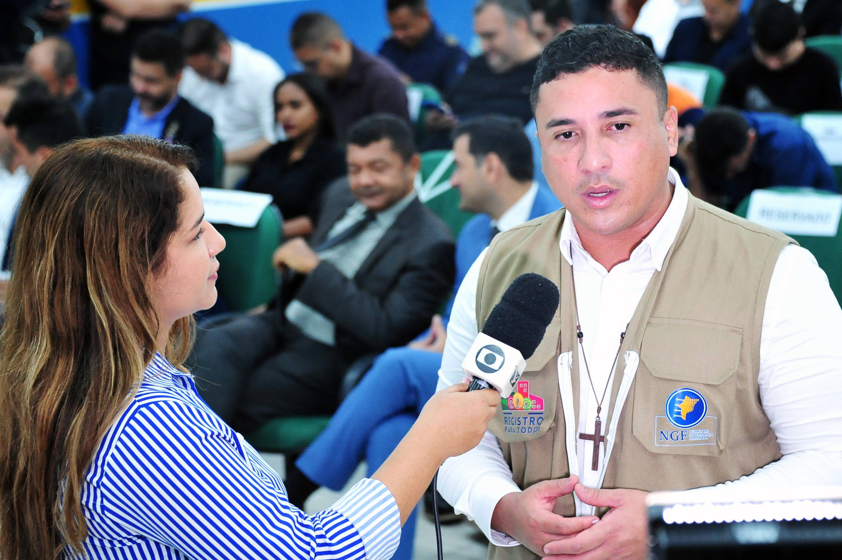 A imagem mostra o coordenador do Núcleo de Governança Fundiária do TJMA, Douglas Lima da Guia, concedendo entrevista a uma repórter. Ele aparece em primeiro plano, usando colete bege com os emblemas do programa ¿Registro para Todos¿ e do NGF. A repórter segura um microfone com a logomarca da TV Mirante/Globo, voltado para ele. Ao fundo, várias pessoas estão sentadas no auditório, entre elas autoridades presentes ao evento. A cena é iluminada, com clima institucional e de cobertura jornalística.
