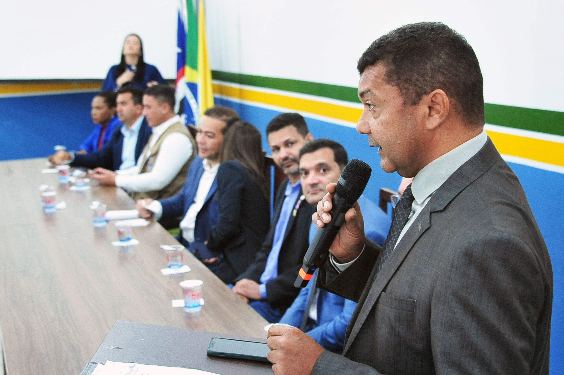 Imagem mostra o juiz Delvan Tavares discursando ao microfone, posicionado à direita da foto, trajando terno cinza. Ele está diante da mesa de autoridades, onde diversos representantes ¿ homens e mulheres ¿ estão sentados, olhando em sua direção. Ao fundo, veem-se bandeiras do Brasil, Maranhão e Imperatriz, além do painel com faixas horizontais nas cores verde, amarela e azul. A cena ocorre no auditório da UAB, com iluminação forte e ambiente institucional.