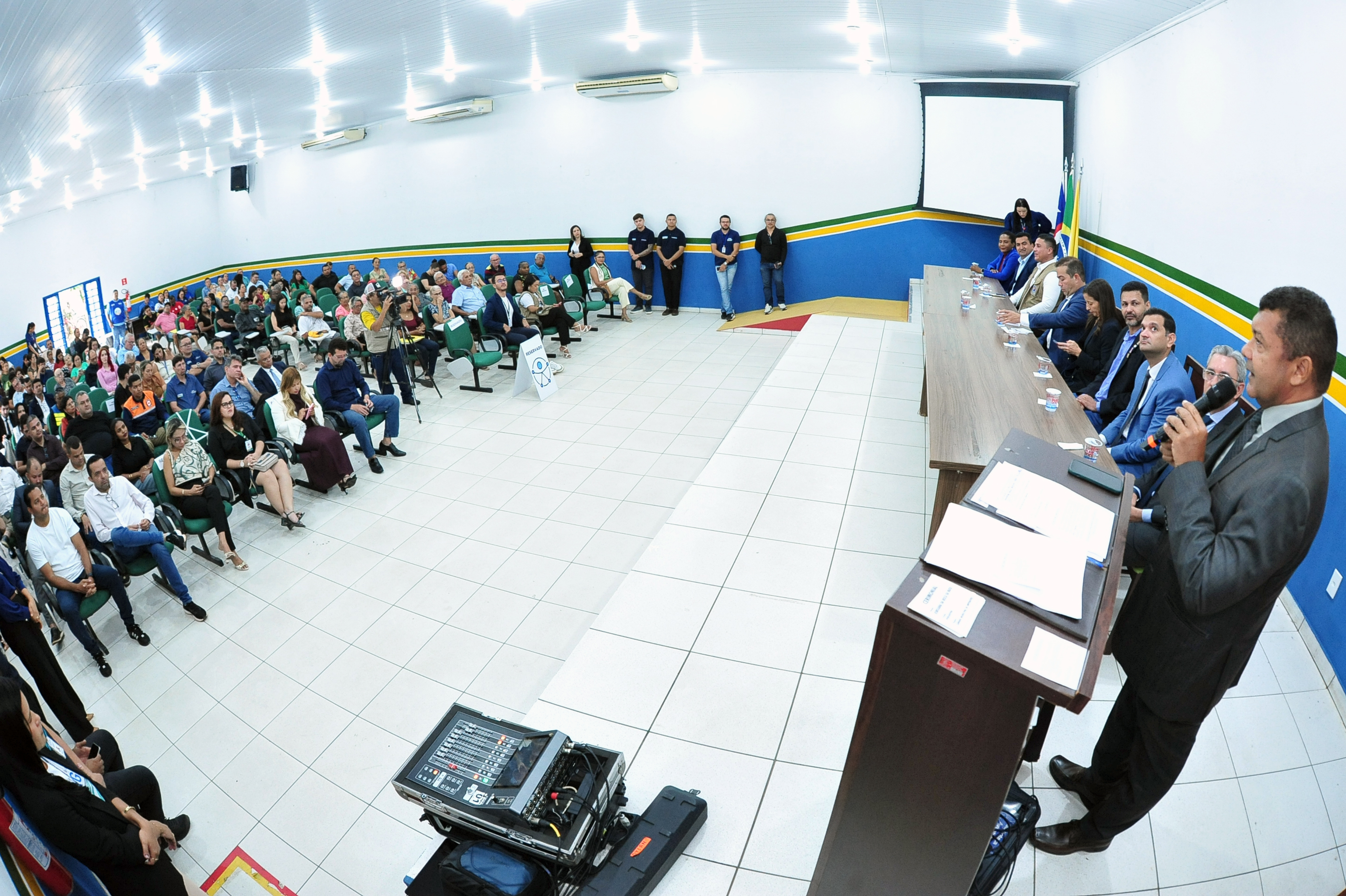 Foto panorâmica do auditório da UAB completamente ocupado. À direita, autoridades estão dispostas à mesa de honra, enquanto o juiz Delvan Tavares fala ao microfone no púlpito. À esquerda, o público ¿ composto por moradores, servidores e representantes de instituições ¿ ocupa as cadeiras verdes. No fundo da imagem, câmeras e equipe técnica registram o evento. O ambiente é amplo, com paredes brancas e faixas em verde, amarelo e azul.