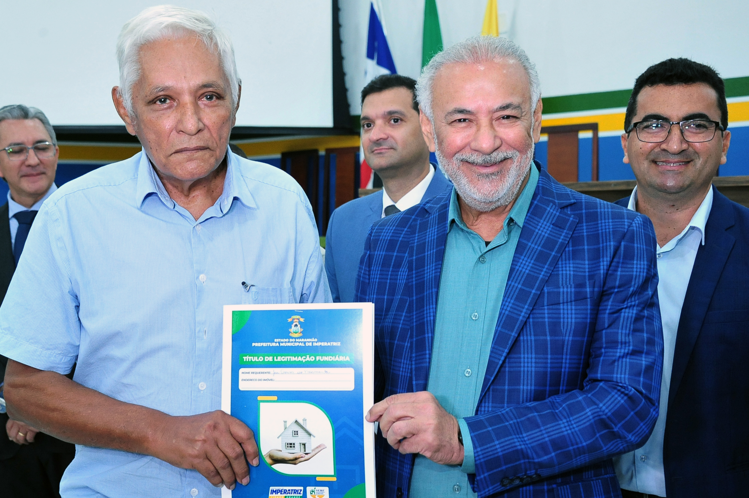 A foto registra um morador idoso, de camisa azul clara, segurando o seu título de legitimação fundiária ao lado do secretário Carlos Hermes, que sorri para a câmera. Ao fundo, outras autoridades acompanham o momento. Bandeiras institucionais aparecem parcialmente à esquerda. O clima é de celebração e entrega formal dos documentos
