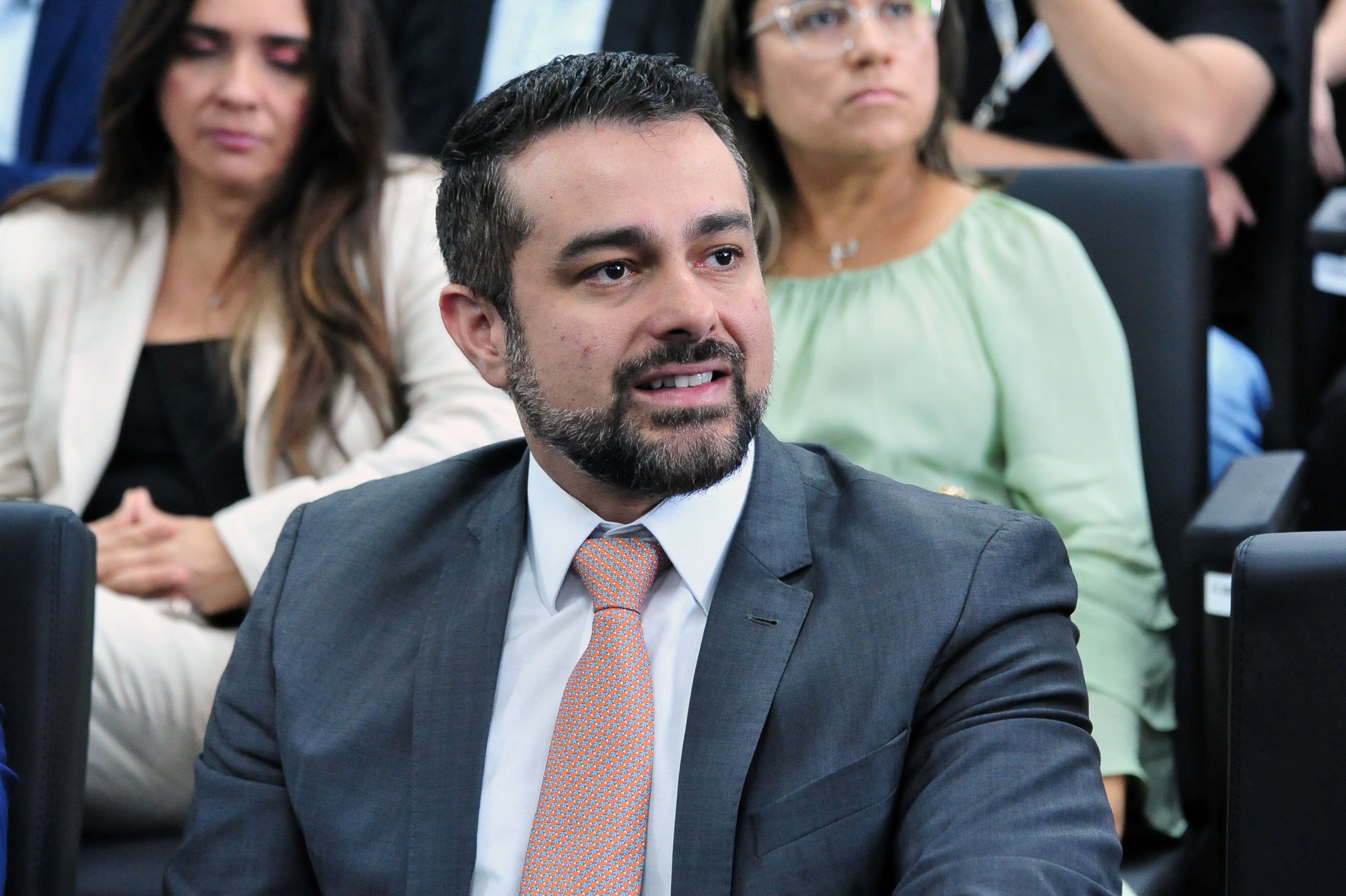 imagem é um close-up que foca em um homem jovem e três pessoas ao seu redor, que estão sentadas na plateia durante um evento formal. ¿¿¿ Pessoa em Foco Homem Central: É um homem jovem, de pele clara e barba bem cuidada (cavanhaque e bigode curtos e escuros).