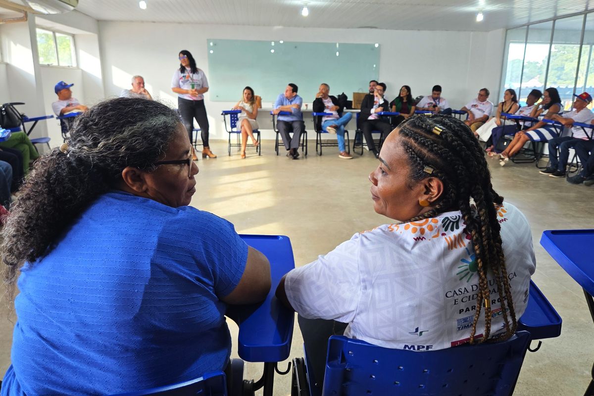 Foto horizontal da Praça da Justiça em Bacabal. Em primeiro plano, destacam-se duas mulheres sentadas em cadeiras azuis, olhando uma para a outra e engajadas em uma conversa. A mulher à esquerda está vestindo uma blusa azul escura. A mulher à direita tem o cabelo penteado em tranças e está usando uma camisa branca com um logomarcas. Em segundo plano, um grupo maior de pessoas está sentado em cadeiras alinhadas, voltadas para a frente da sala. No centro, uma mulher está de pé, falando para o grupo, próxima a um quadro branco. Os participantes no fundo estão vestidos de maneira casual.