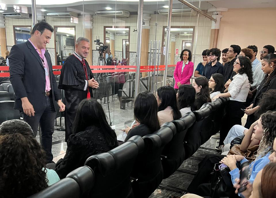 A imagem registra o desembargador Ronaldo Maciel, de toga, conversando com estudantes que estão sentados nas cadeiras fronteiras ao Plenário. Um servidor de camisa rosa e blazer escuro está ao lado do magistrado. Atrás do grupo, há paredes de vidro que dão vista para o Plenário, onde é possível ver câmeras e equipamentos da sessão em andamento.