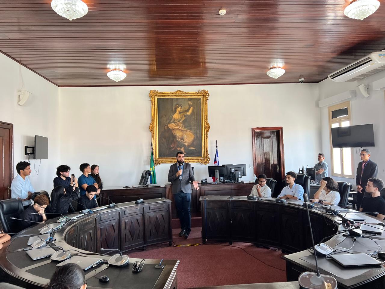 Imagem mostra o Salão do Pleno do Tribunal de Justiça do Maranhão. Estudantes estão sentados ao redor da mesa semicircular, enquanto um servidor ¿ em pé, ao centro, usando terno cinza ¿ fala ao grupo. No fundo, há um grande quadro clássico em moldura dourada e bandeiras posicionadas atrás da mesa principal. A iluminação é feita por lustres e o teto é de madeira.