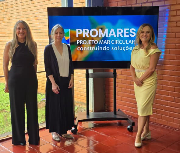 Três mulheres posam sorrindo diante de uma tela grande que exibe o logotipo do Projeto PROMARES ¿ ¿Mar Circular: Construindo Soluções¿. À esquerda, uma mulher veste blusa preta sem mangas e calça preta. Ao centro, outra mulher usa cardigã escuro, blusa clara e saia longa azul-marinho. À direita, a juíza Teresa Mendes aparece com vestido amarelo claro, segurando uma bolsa, também sorrindo. O fundo é composto por parede de tijolos aparentes e piso vermelho.