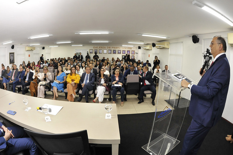 Uma sala ampla com diversas pessoas sentadas em cadeiras. À frente, à direita, um homem de terno escuro e gravata vermelha fala ao microfone em um púlpito de acrílico transparente. Ele está virado para o público, que inclui homens e mulheres vestidos formalmente. No fundo, há quadros e uma faixa com a frase "Galeria de Presidentes"
