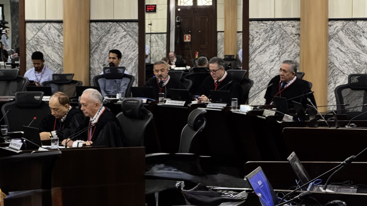 Foto horizontal de desembargadores do TJMA durante sessão do Órgão Especial. A imagem mostra a sala de sessões plenárias do Tribunal. Há várias pessoas sentadas em cadeiras de escritório, algumas vestindo togas pretas com detalhes vermelhos. As mesas estão equipadas com laptops, microfones e copos de água. Ao fundo, há uma porta de madeira e paredes revestidas de mármore. Há também uma tela digital com números vermelhos no canto superior esquerdo da imagem.