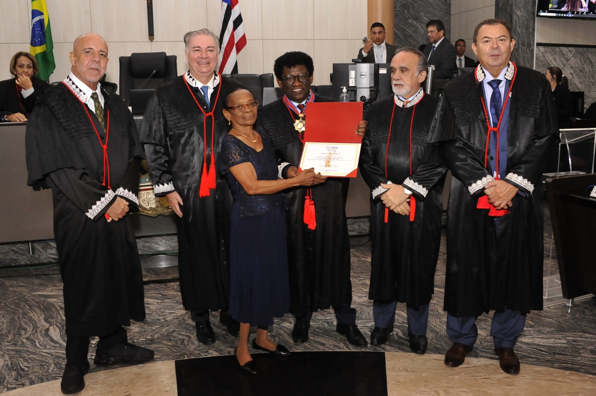 Foto horizontal da diplomação do desembargador Raimundo Neris, com integrantes da mesa diretora do TJMA e dona Neide, mãe do homenageado. A imagem mostra um grupo de seis pessoas em pé, vestidas com trajes formais, de togas jurídicas, com detalhes em vermelho e branco. O desembargador homenageado, no centro, está segurando o certificado, que está sendo entregue por uma mulher ao lado, que é sua mãe. Ao fundo, há bandeiras do Brasil e do Maranhão Maranhão. A cena é  uma cerimônia na sala das sessões plenárias do TJMA.