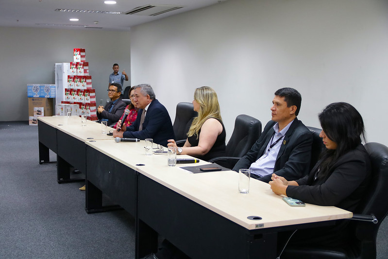 Foto horizontal Encontro final de 2024 da DSI  e Comissão Permanente de Segurança Institucional no Fórum de São Luís. A imagem a mesa longa de autoridades no auditório do Fórum de São Luís, onde estão sentadas seis pessoas, entre elas o desembargador Bonfim, que fala ao microfone. Além do microfone, a mesa tem copos de água e alguns documentos. Ao fundo, há uma pilha de caixas empilhadas e um refrigerador. Uma pessoa está em pé ao fundo, aparentemente observando a reunião. A sala é bem iluminada em um ambiente formal.