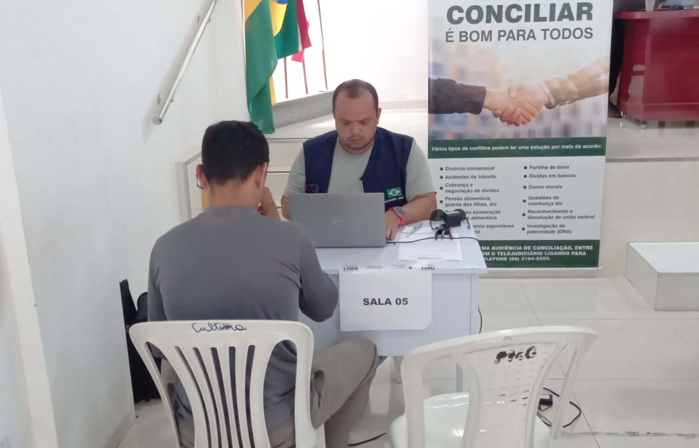 Foto colorida mostra conciliador atendendo um cidadão durante o projeto Conciliação Itinerante em Grajaú. Em frente ao servidor existe um computador. Atrás deles, existe um banner do projeto.