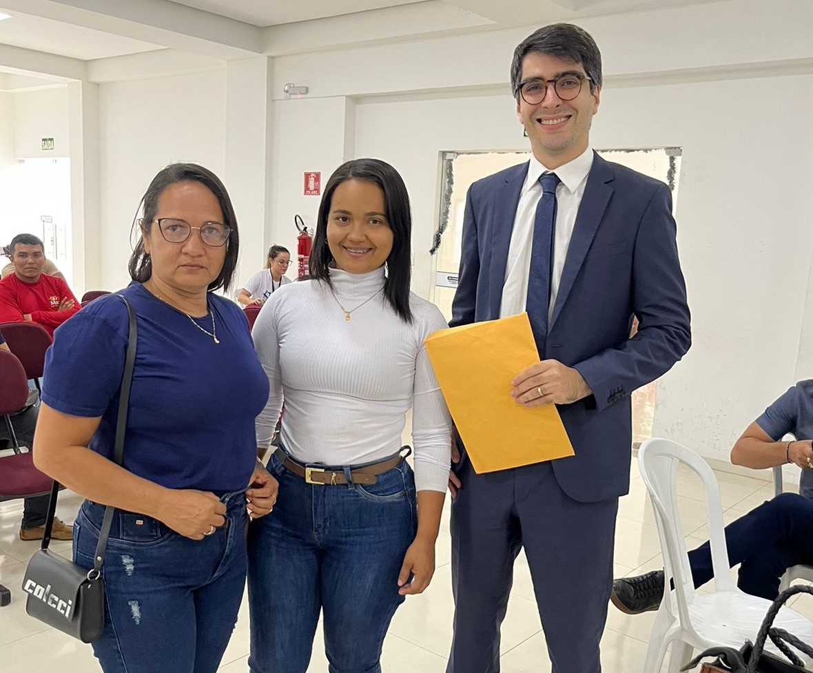Foto colorida mostra duas mulheres em pé ao lado do juiz Bernardo Freire, que segura um envelope, durante evento Conciliação Itinerante em Grajaú.