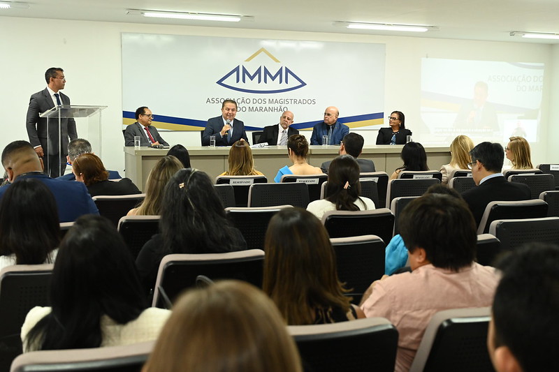 Foto horizontal da abertura do Seminário de Saúde do TJMA. A imagem mostra um auditório. Na frente da sala, há uma mesa com cinco pessoas sentadas, todas vestidas formalmente. Uma pessoa está de pé ao lado de um púlpito. Atrás da mesa, há um grande painel com o logotipo e o nome "Associação dos Magistrados do Maranhão (AMMA)". A audiência está sentada em cadeiras, prestando atenção aos palestrantes. Há também uma tela de projeção à direita, mostrando uma imagem ampliada de uma das pessoas na mesa.

Esta imagem retrata um evento formal, possivelmente uma conferência ou reunião importante, envolvendo magistrados ou juízes do estado do Maranhão, no Brasil.