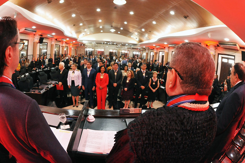 Foto horizontal da Sessão Solene de 211 anos do TJMA com entrega de medalhas. A imagem mostra a cerimônia formal na Sala das Sessões Plenárias do Tribunal, um salão bem iluminado. Há várias pessoas de pé, vestidas formalmente, algumas usando medalhas. A perspectiva é do ponto de vista de uma pessoa no alto, olhando para a plateia. No centro há uma mesa com documentos e um microfone. A palavra "PRESIDENTE" está visível na frente da mesa. O salão tem um teto curvo com iluminação embutida e luzes vermelhas nas laterais. A audiência está atenta e respeitosa.