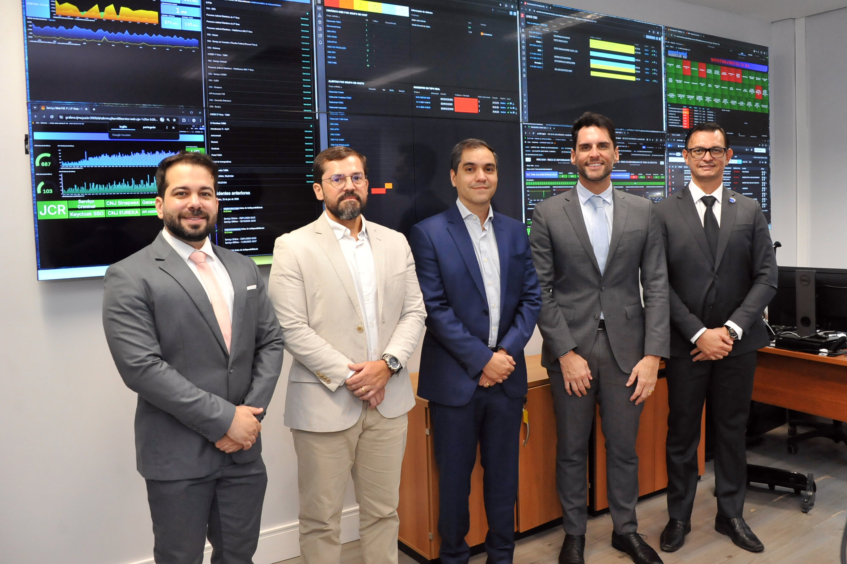 TJMA: Portal do Poder Judiciário do Estado do Maranhão 2 Em um ambiente interno e bem iluminado, cinco homens adultos, de tons de pele variados (entre brancos e pardos), estão em pé, lado a lado. Todos vestem trajes formais; quatro deles usam ternos em tons de cinza e azul, enquanto o segundo da esquerda para a direita veste um blazer bege sobre camisa branca. Eles sorriem levemente, transmitindo uma postura profissional e cordial. Ao fundo, a parede é ocupada por grandes monitores de vídeo (videowall) repletos de dados técnicos, gráficos de desempenho e tabelas com fundo preto e fontes coloridas (verde, azul e amarelo), indicando um centro de controle ou núcleo de TI. À direita, vê-se parte de uma mesa de escritório de madeira com um monitor de computador.