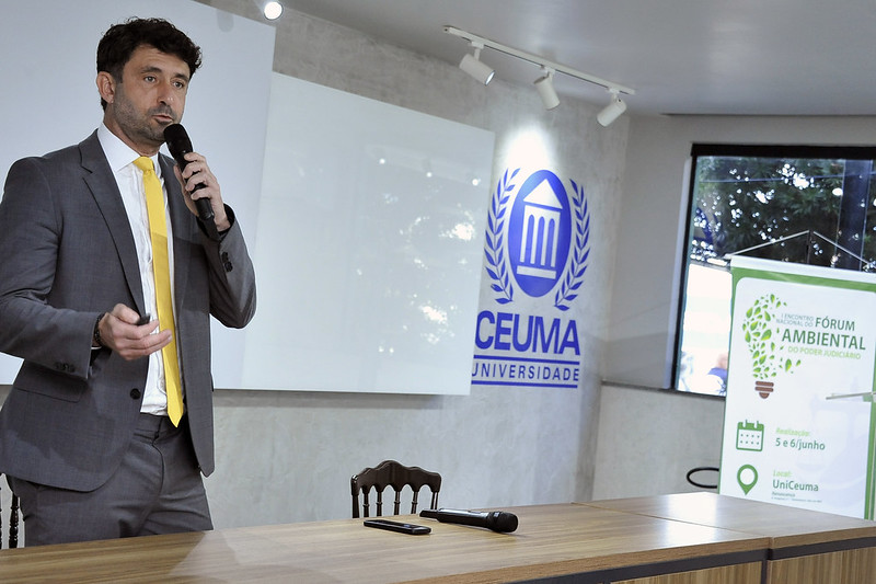 A imagem mostra um homem em pé à esquerda de uma mesa de madeira, segurando um microfone na mão direita e gesticulando com a esquerda. Ele veste um terno cinza e uma gravata amarela.  Ao fundo, há uma parede clara com o logotipo azul da "CEUMA UNIVERSIDADE", que inclui um emblema com folhas de louro e um chapéu de formatura. À direita do logotipo da universidade, há um banner verde que, embora parcialmente visível, exibe o título "ENCONTRO NACIONAL DE DIREITO AMBIENTAL DO PODER JUDICIÁRIO". O banner também menciona "5 e 6/JUNHO" como datas de realização e "Local: UniCeuma".  Sobre a mesa de madeira, há alguns objetos, incluindo o que parecem ser canetas e um microfone extra. A iluminação ambiente é feita por alguns focos de luz no teto.