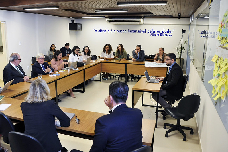 A imagem mostra uma sala de reunião com várias pessoas sentadas em torno de uma grande mesa em formato de "U" ou retangular, criando um ambiente de colaboração.  No fundo da sala, há uma parede branca com a frase "A ciência é a busca incansável pela verdade." atribuída a Albert Einstein, escrita em letras azuis. No teto, há um forro de madeira com iluminação embutida.  As pessoas na mesa estão vestidas de forma formal ou semi-formal, e a maioria delas está interagindo, algumas usando notebooks abertos, indicando que a reunião é produtiva. Há aproximadamente 15 pessoas visíveis na imagem.  No lado direito da imagem, há uma parede de vidro, e uma lousa de vidro no canto direito tem vários post-its amarelos e verdes, sugerindo brainstorming ou organização de ideias.  A iluminação da sala é clara e uniforme, contribuindo para a atmosfera de trabalho