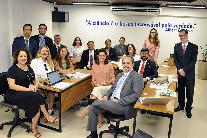 A imagem retrata um grupo de aproximadamente 15 pessoas, sentadas e em pé, em um ambiente que se assemelha a uma sala de reuniões ou de treinamento.  A maioria das pessoas está sentada em torno de mesas claras, algumas com laptops abertos e documentos espalhados, o que sugere um contexto de trabalho ou aprendizado. Há uma mistura de homens e mulheres, predominantemente em trajes formais ou semi-formais.  No fundo da sala, há uma parede branca com a citação em azul: "A ciência é a busca incansável pela verdade." atribuída a Albert Einstein. No teto, há um forro de madeira com luminárias.  A iluminação é clara e uniforme, e a atmosfera geral parece ser de profissionalismo e colaboração. Algumas pessoas olham diretamente para a câmera, indicando que a foto foi posada.
